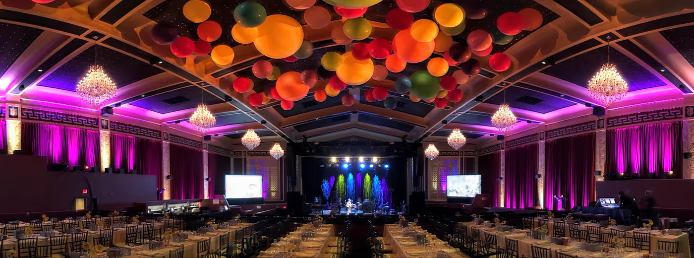 Large banquet hall decorated with purple curtains, chandeliers, and colorful balloons hanging from the ceiling. Multiple tables are set with plates, glasses, and napkins, arranged in front of a stage with bright lights and a band performing.