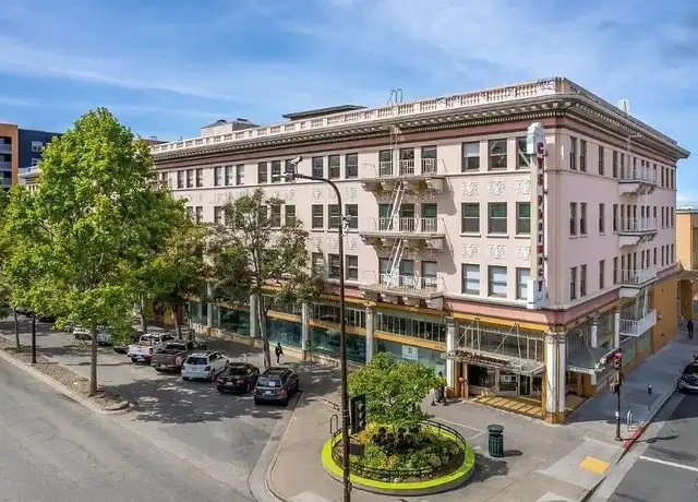 Street view of a corner building with a cafe or restaurant on the ground floor, parked cars along the street, and trees in a downtown area.