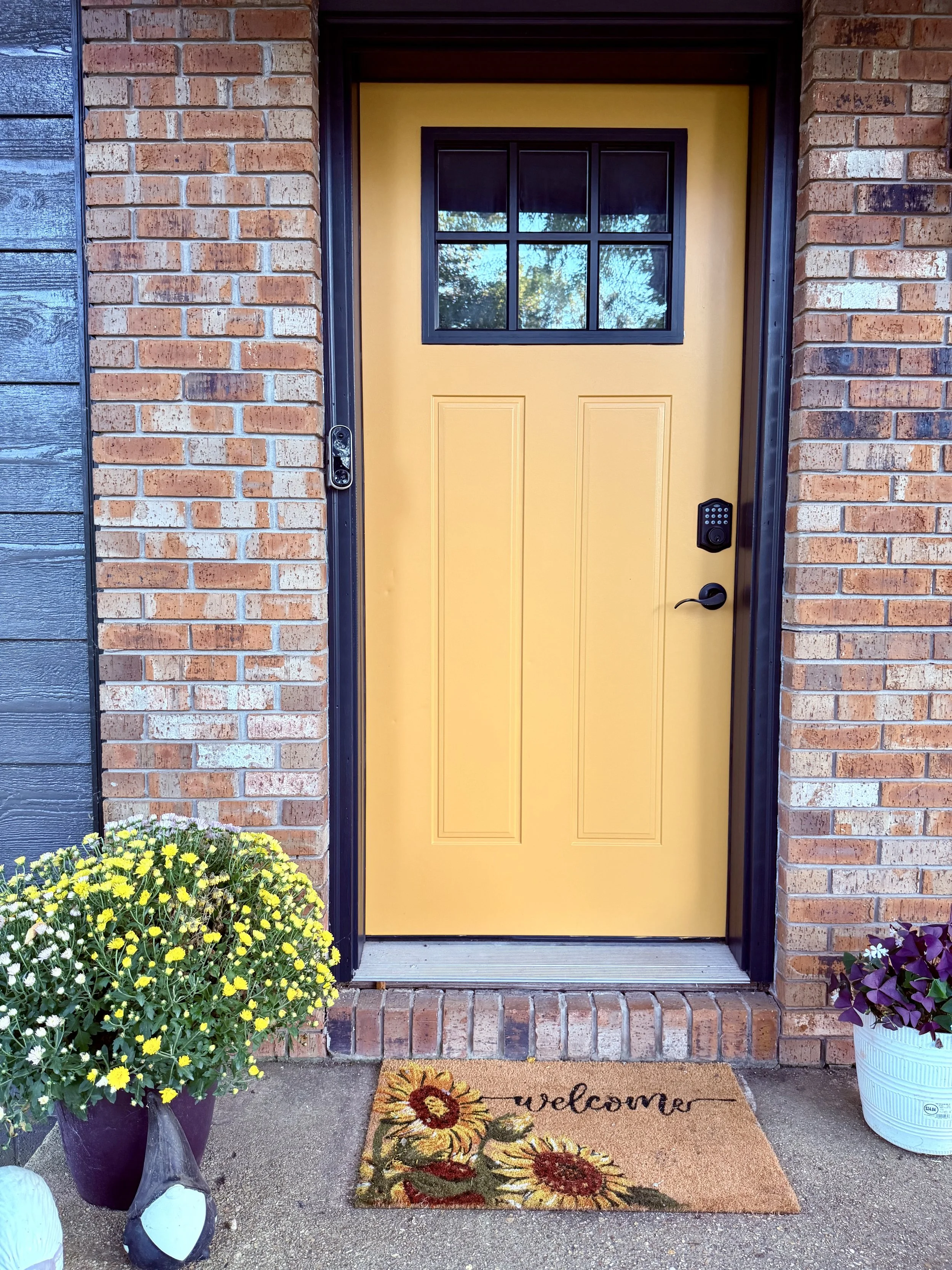Front door with a yellow painted door, black trim, and a small window at the top. There is a welcome mat with sunflower designs on the doorstep. Potted yellow flowers are on the left side and purple flowers on the right side.