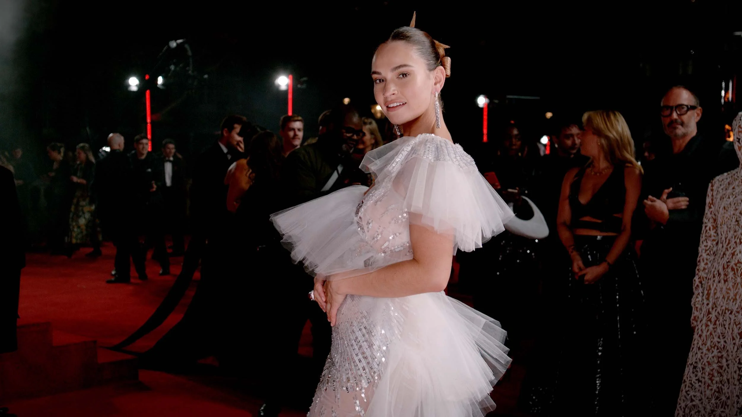 A woman in a white wedding dress with glambot, details is at a formal event, surrounded by other guests in evening attire. She is smiling and looking at the camera, with her hair styled in an elegant updo.