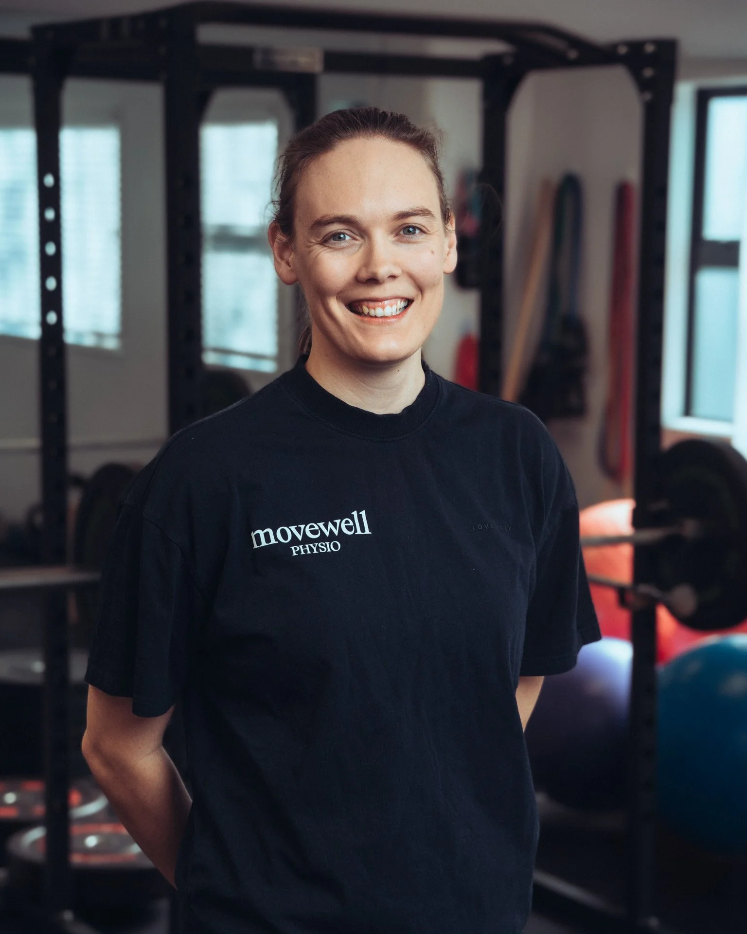 A smiling woman with brown hair pulled back, wearing a black shirt with white text that reads 'movewell PHYSIO', standing in a gym with workout equipment and large windows in the background.