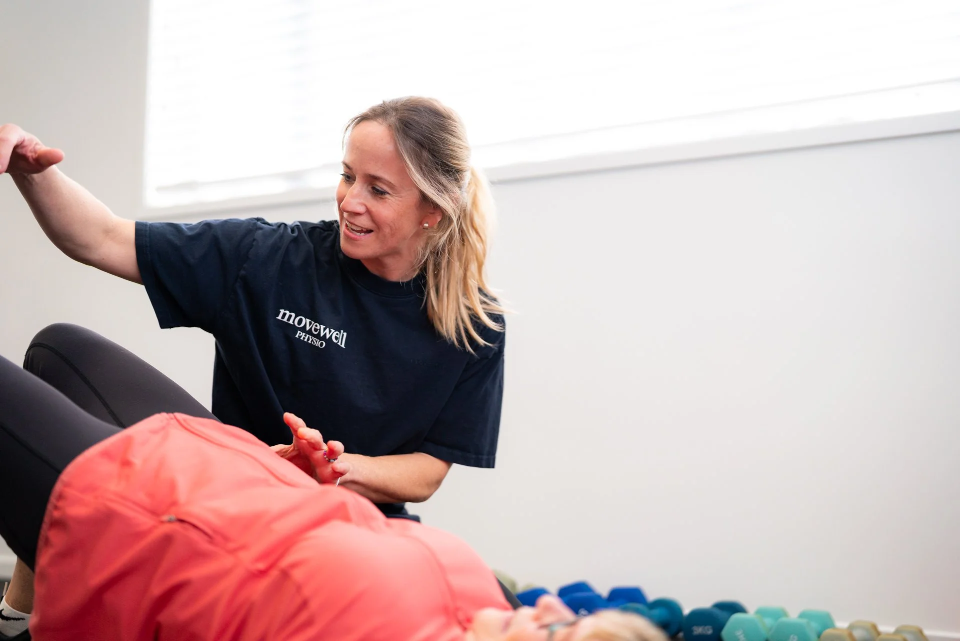 A woman in a dark T-shirt with 'movewell PHYSIO' printed on it is smiling and helping a person lying on a massage table. The person is wearing a coral-colored jacket and has their hands clasped, with colorful dumbbells on the floor near them. Bright natural light comes through a window with blinds in the background.