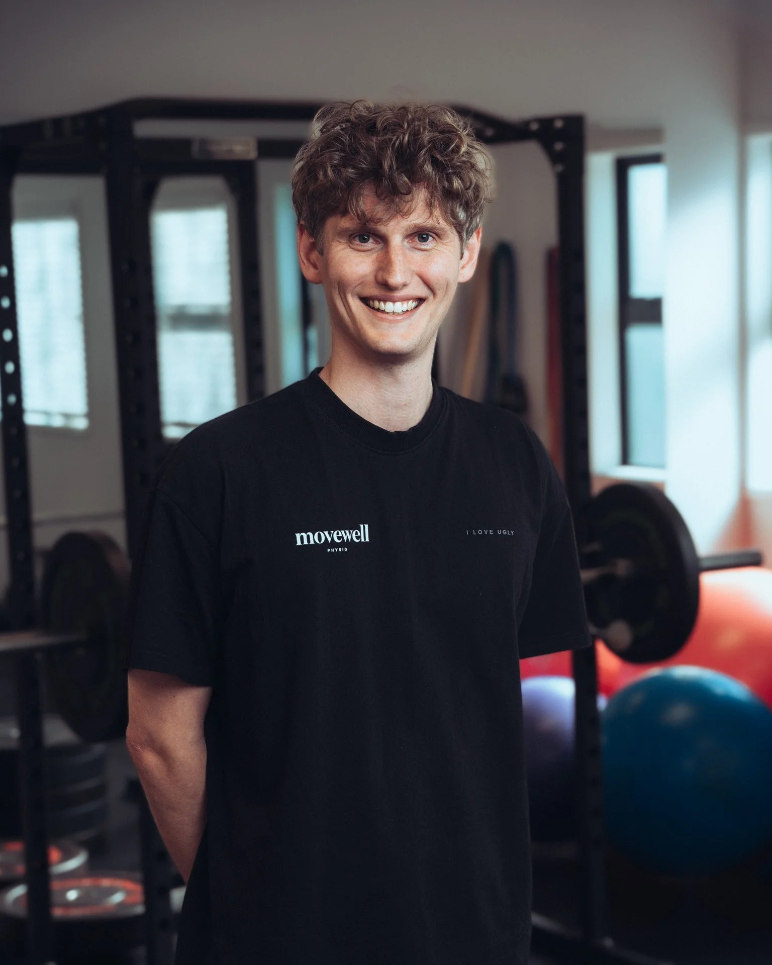 A smiling young man with curly hair standing in a gym, wearing a black t-shirt with the words 'movewell' and 'I LOVE UGLY' printed on it.