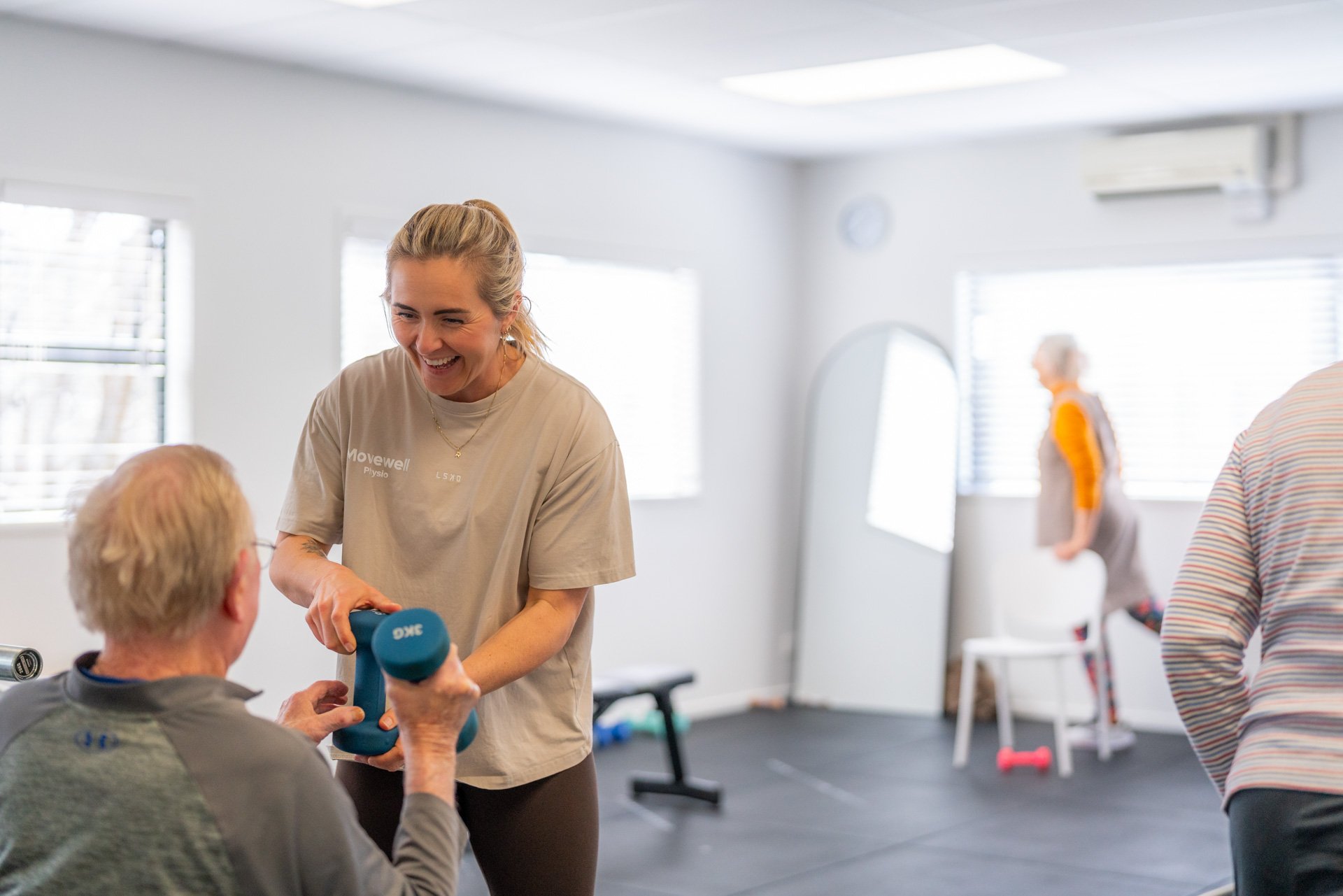 A female fitness instructor demonstrating weightlifting to an elderly man during exercise class in a bright gym.