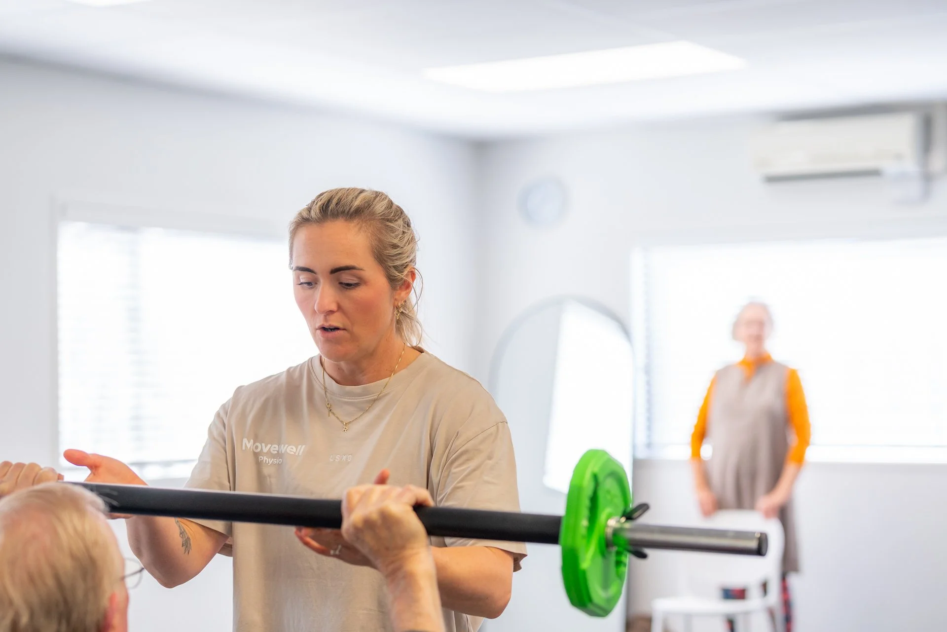 A woman with gray hair tied back working with an elderly person's arm in a physical therapy or rehabilitation setting. Another woman is in the background near a window, wearing a gray apron with orange sleeves, standing in a bright room with white walls and large windows.
