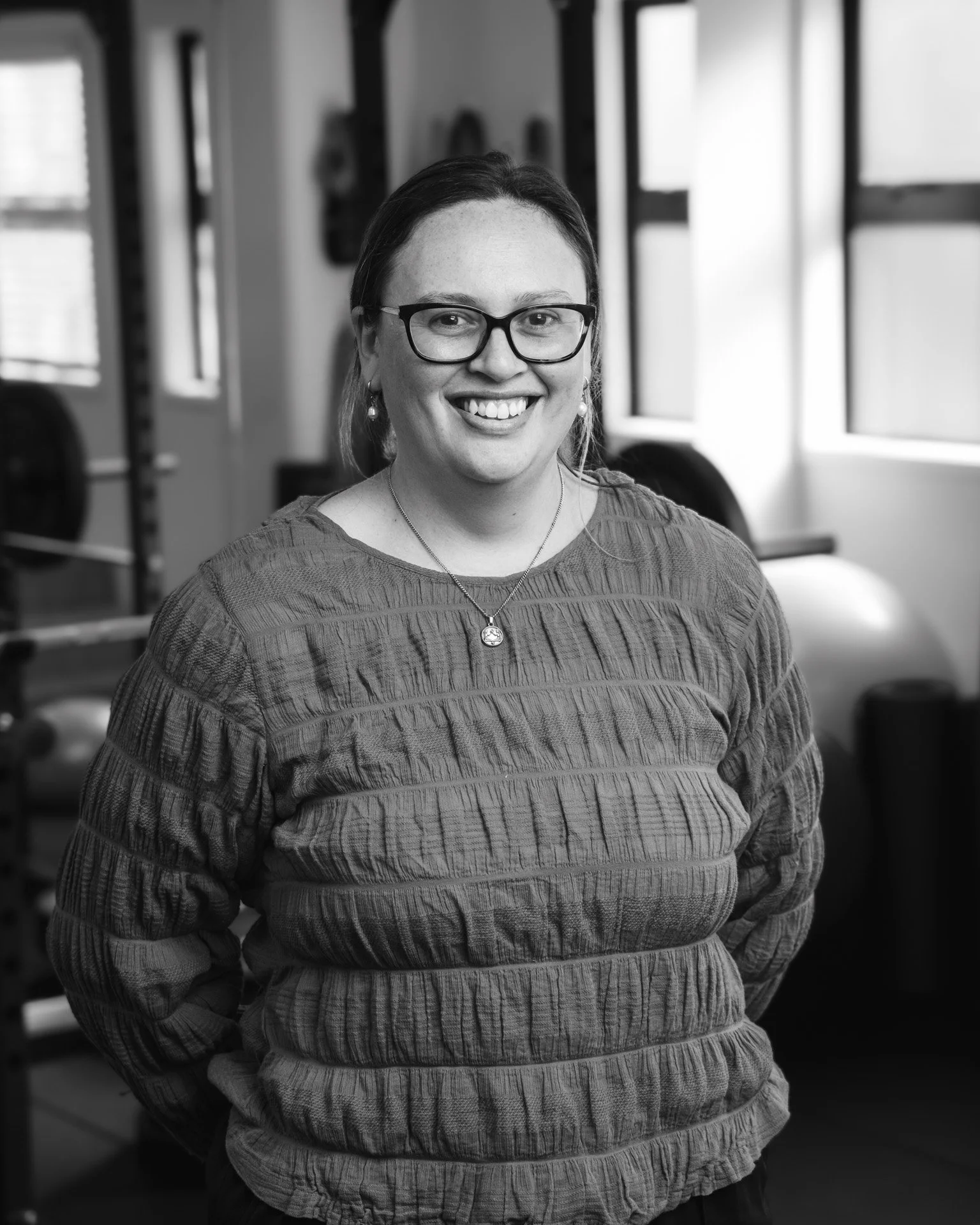 Black and white photo of a smiling woman with glasses, wearing a striped sweater, standing in a room with windows and exercise equipment in the background.