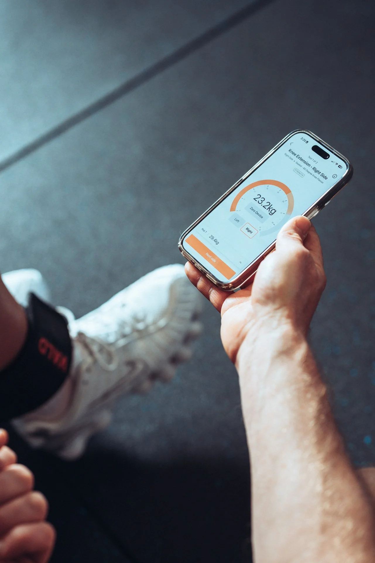 A person holding a smartphone displaying a weight tracking app with a current weight of 23.2 kg, showing a progress chart and options for left or right knee measurement, and a button labeled 'Next Side'.