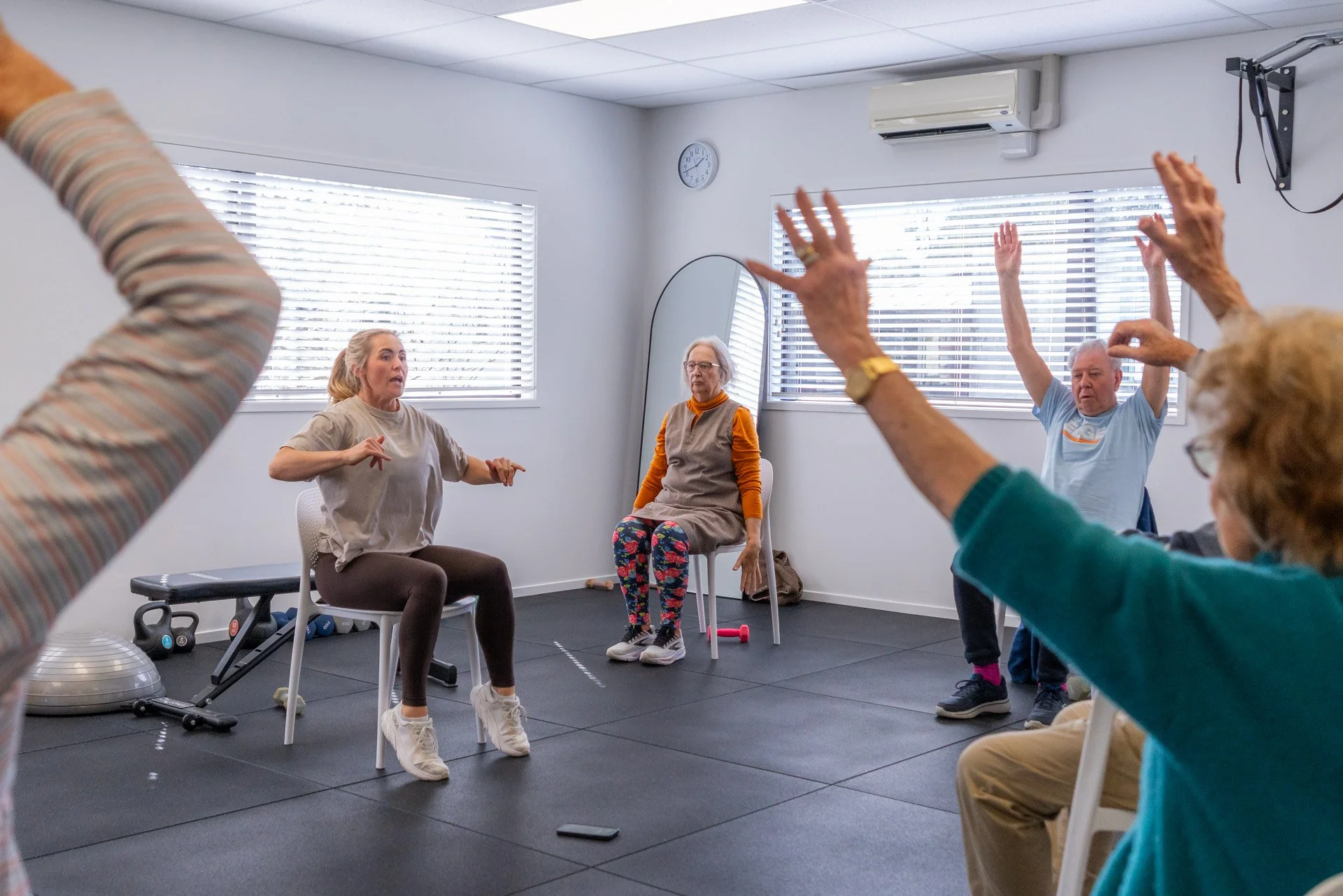 A group of seniors participating in a seated exercise class, with some raising their hands as directed by the instructor. The room has large windows with blinds, a wall clock, and exercise equipment in the background.