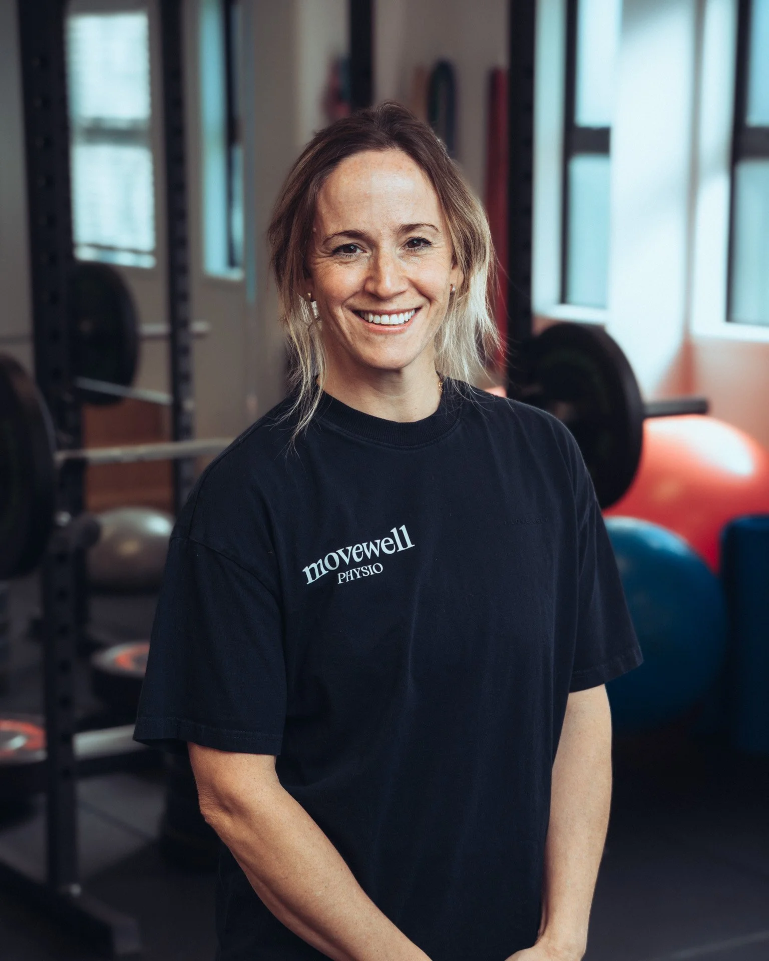 A smiling woman with shoulder-length light brown hair, wearing a black t-shirt with the words 'movewell PHYSIO' on it, inside a gym with weightlifting equipment and large windows.