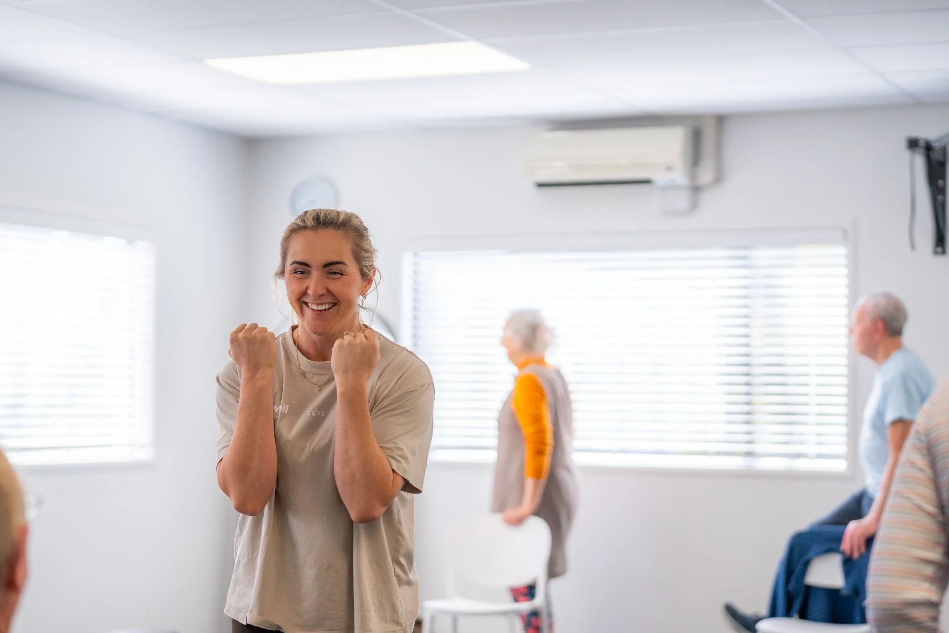 Woman in beige t-shirt celebrating with clenched fists in a bright room, with elderly people sitting and walking in the background.