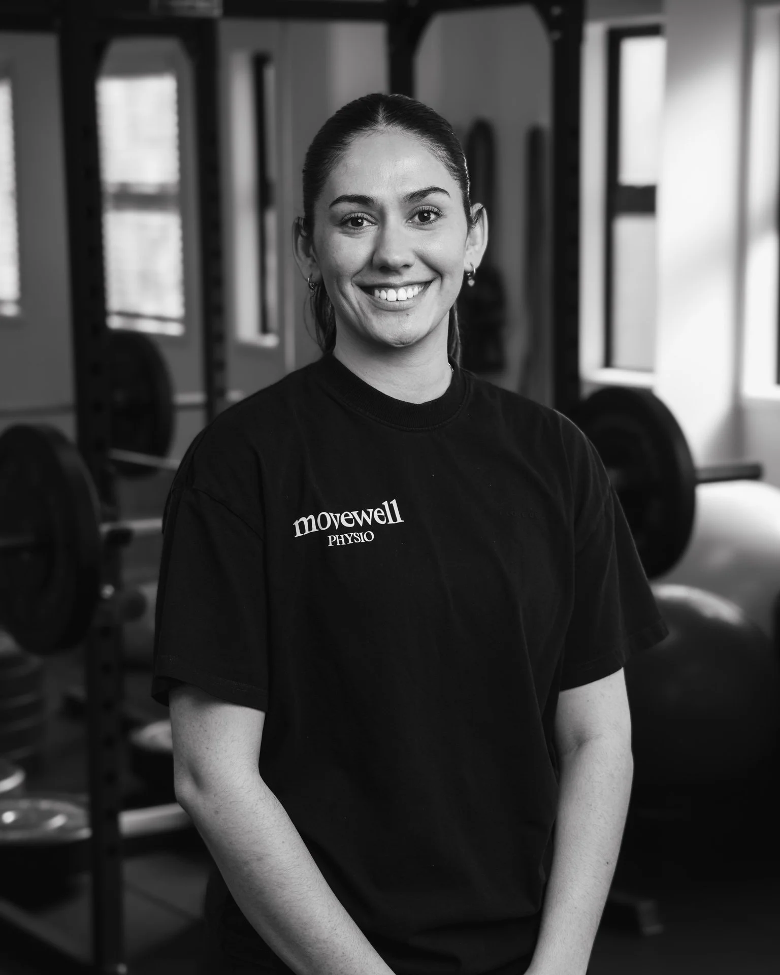 A smiling woman with dark hair tied back, wearing a black t-shirt with the logo 'movewell PHYSIO,' standing in a gym or fitness studio with weightlifting equipment in the background.