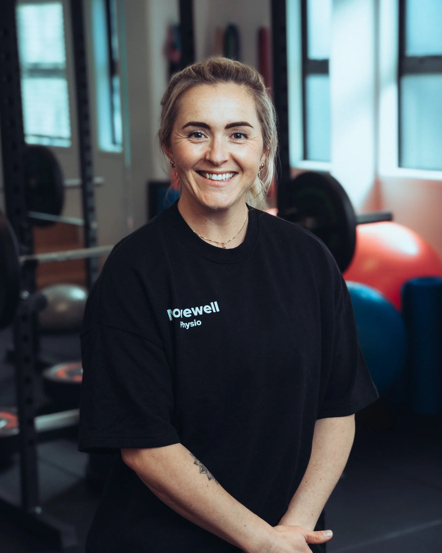 A smiling woman with blonde hair tied back, wearing a black shirt with white text, standing inside a gym with fitness equipment and large windows in the background.