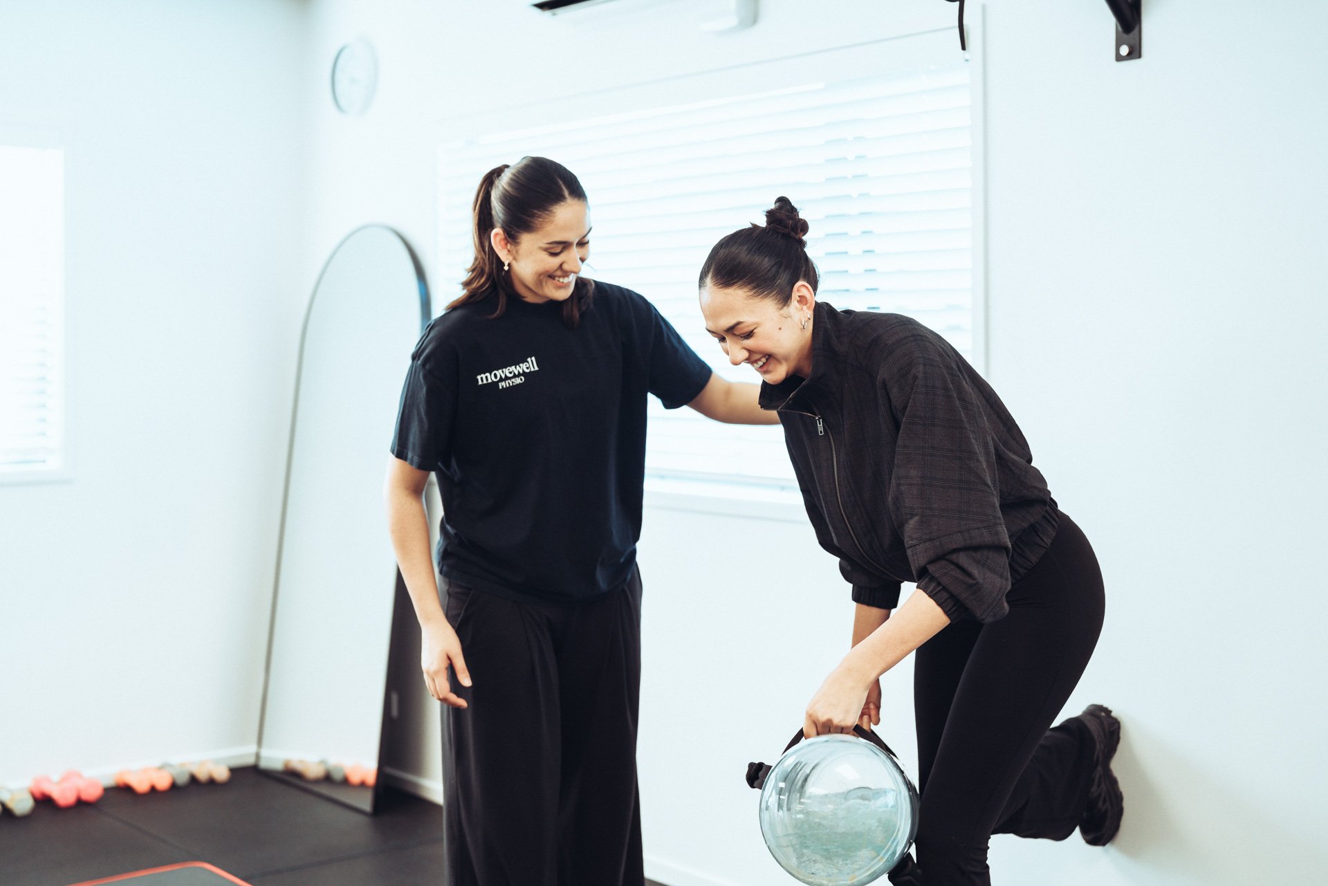 A woman in a black Moveswell Physiotherapy shirt assisting another woman with a knee exercise in a bright, minimalistic fitness room. The woman on the floor is holding a glass exercise ball.