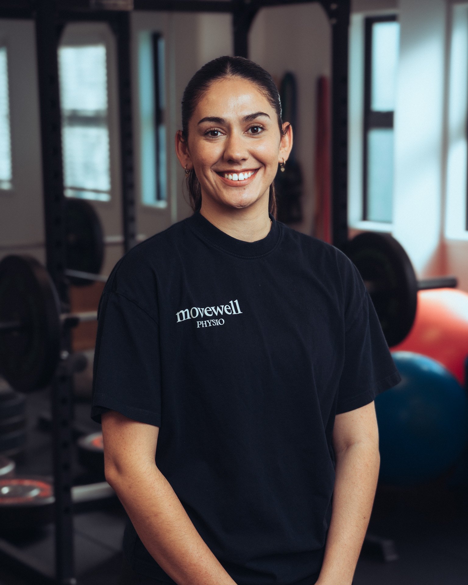 Smiling woman with dark hair in a bun, wearing a black t-shirt with 'movewell PHYSIO' logo, standing in a gym with weights and workout equipment in the background.