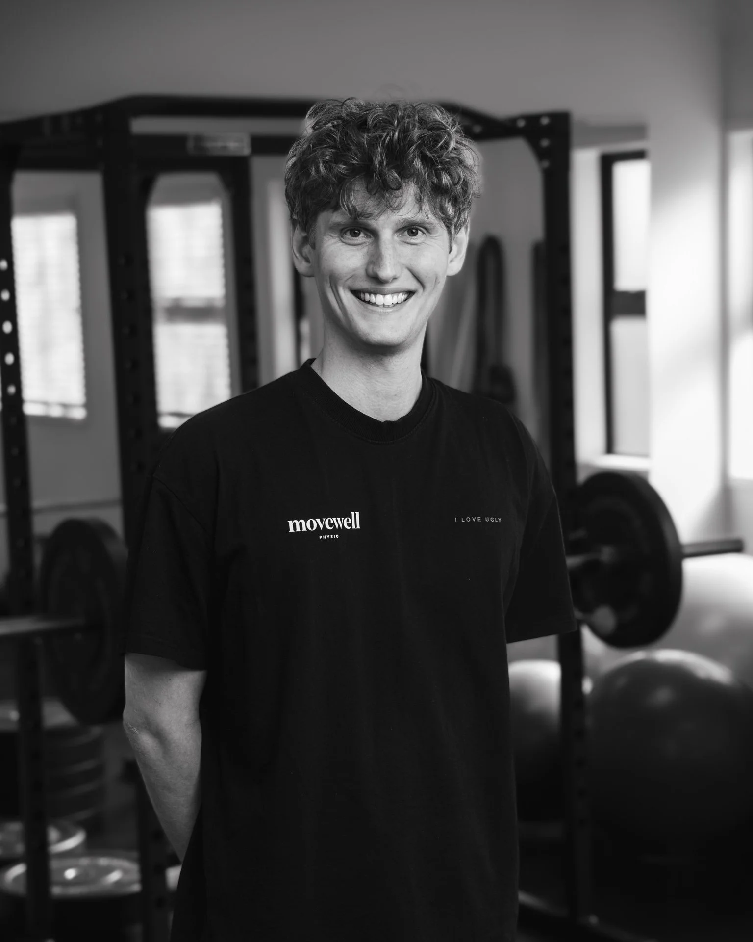 A young man with curly hair smiling in a gym, standing in front of weightlifting equipment, wearing a black t-shirt with text on it.