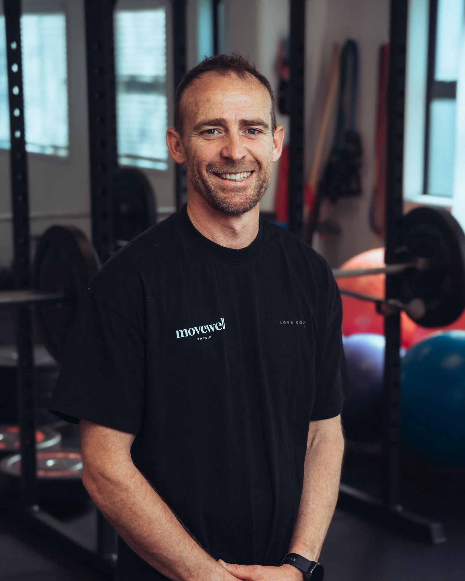 A smiling man with short dark hair, wearing a black T-shirt and a watch, stands in a gym with weightlifting equipment, barbells, and weights visible in the background.