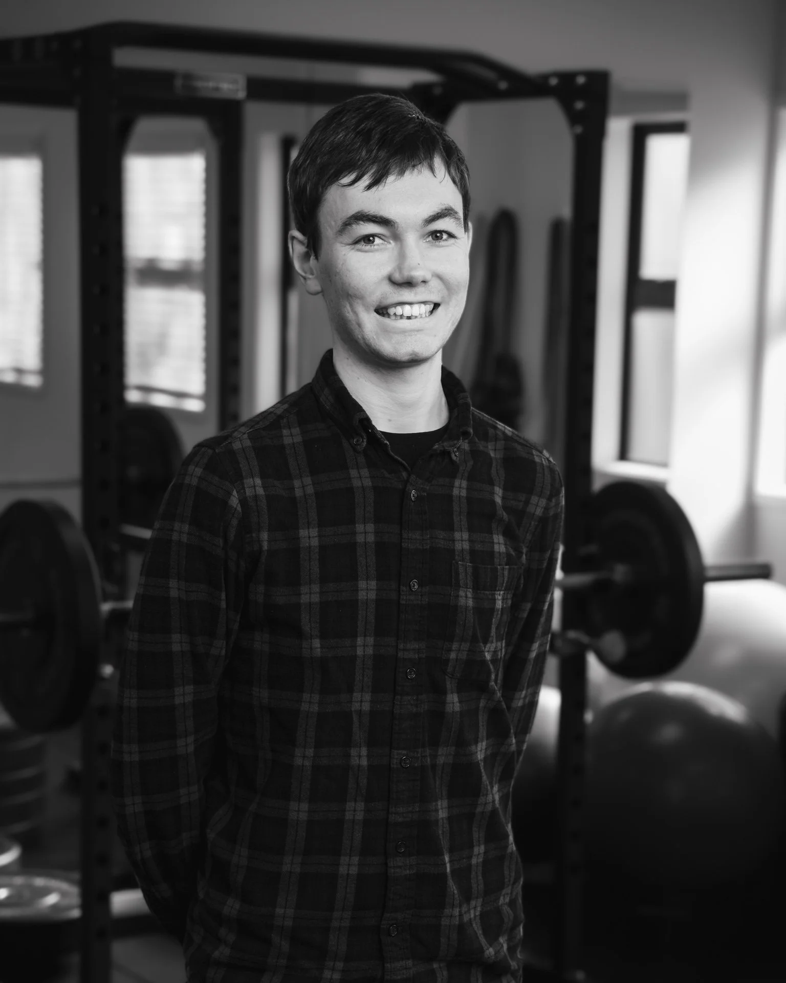 A young man smiling, standing in a gym with weightlifting equipment in the background.