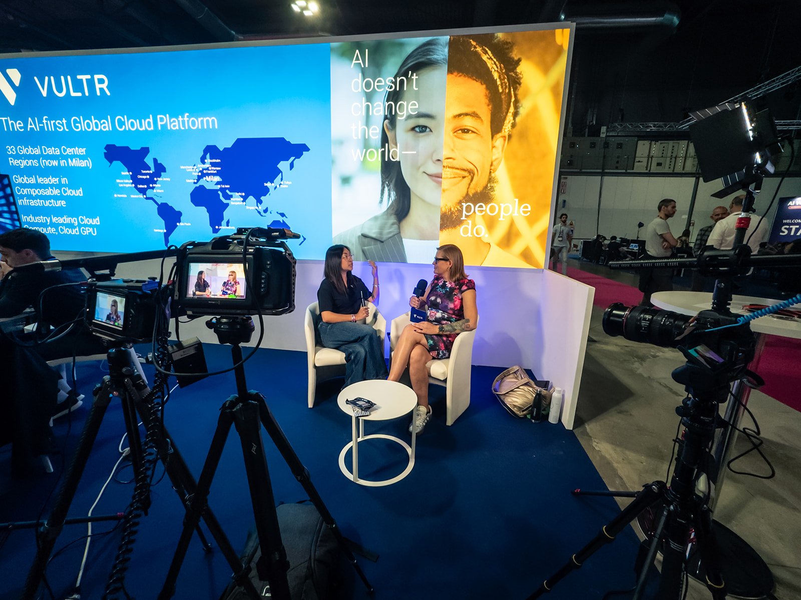 Two women are sitting and having an interview in front of a large screen with a blue and orange graphic. The screen displays the VULTR logo, a world map, and the text 'The AI-first Global Cloud Platform' and 'AI doesn't change the world—people do.' Cameras and lighting equipment are set up around them, and several people are visible in the background at an industry event.