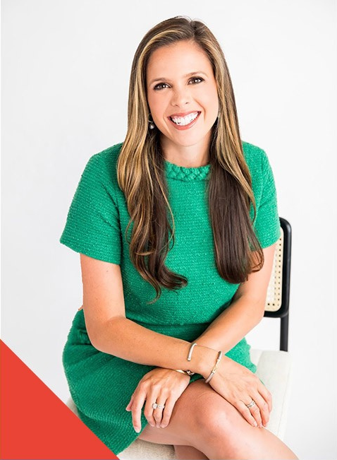 A young woman with long brown hair, wearing a green dress, sitting on a white chair, smiling at the camera against a plain white background.