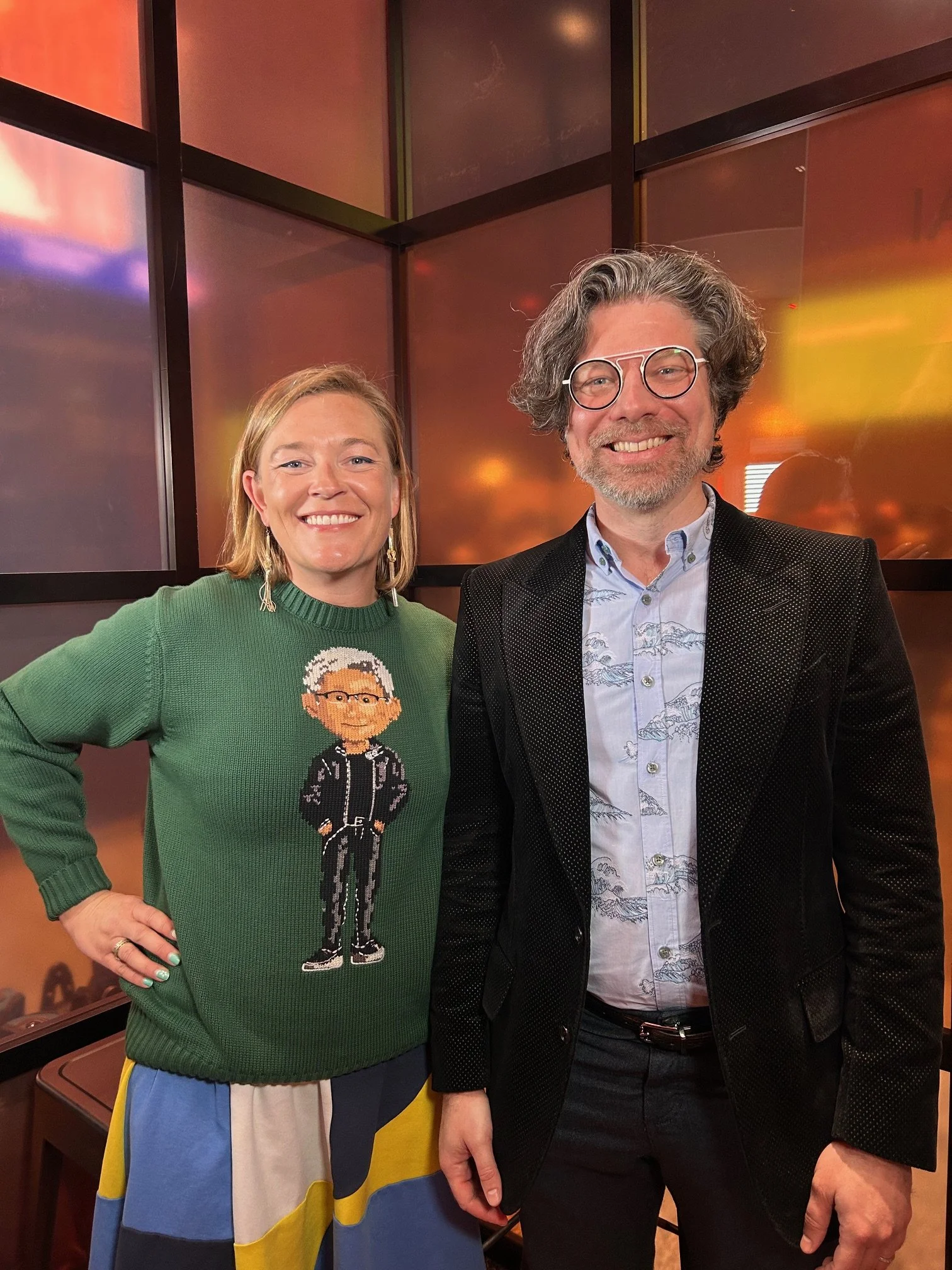 Two smiling individuals, a woman on the left wearing a green sweater with an illustrated character and a man on the right wearing glasses, a patterned shirt, and a black blazer, standing indoors with a colorful, reflective background.