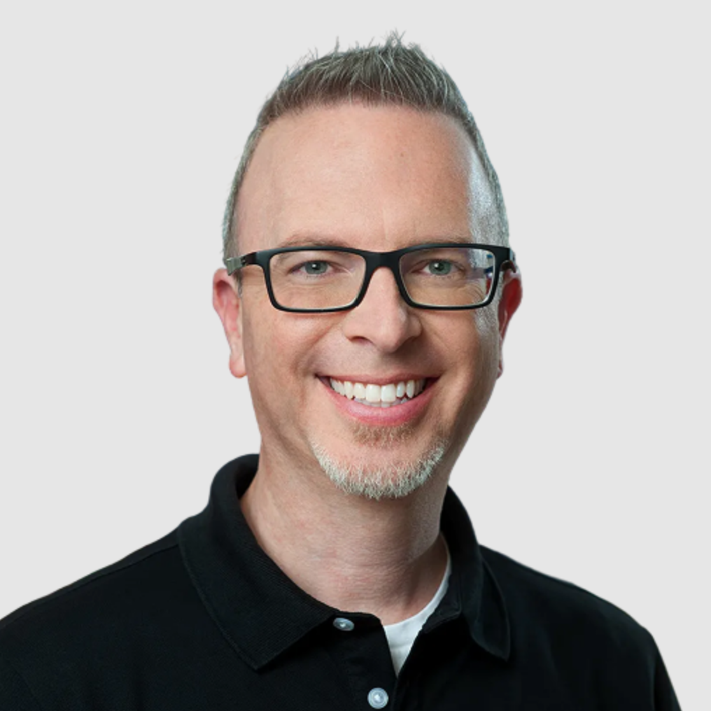 Headshot of a smiling man with glasses and light-colored hair styled upward, wearing a black collared shirt against a plain background.