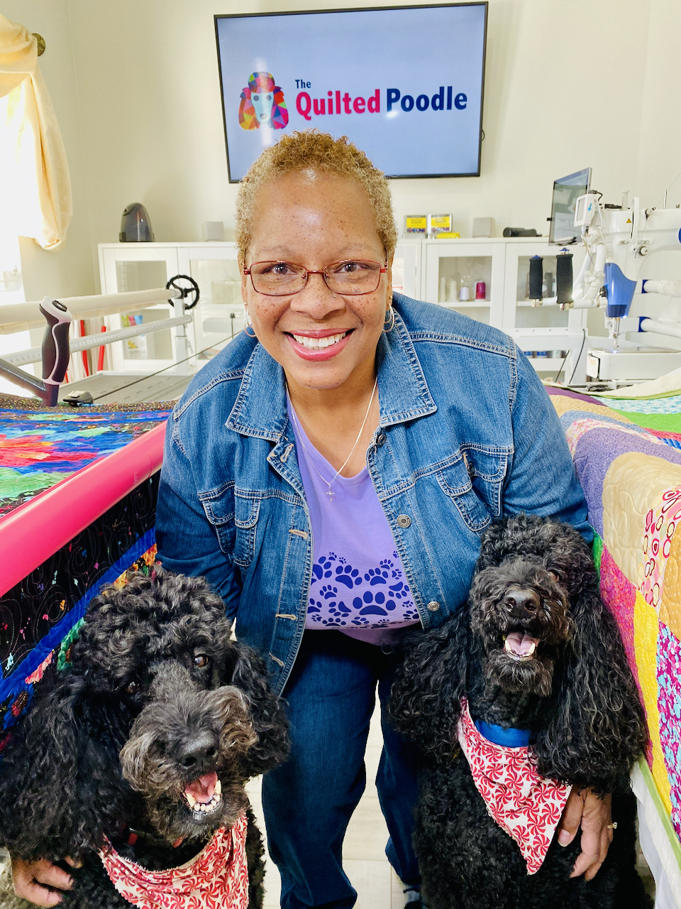 Smiling woman in glasses and denim jacket posing with two black Poodle dogs, in a room with quilting supplies and a TV screen that reads 'The Quilted Poodle'.
