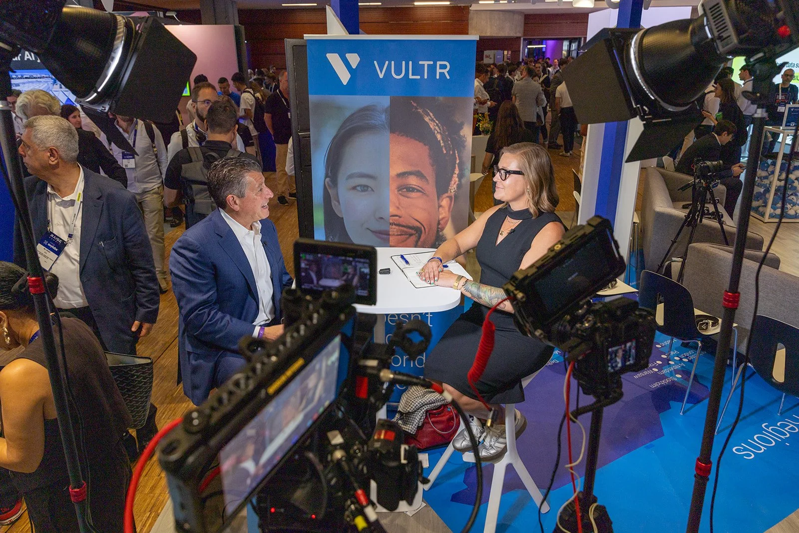 An interview or discussion taking place at a conference or event, with two people seated at a table surrounded by cameras and lighting equipment, and a large VULTR banner in the background.