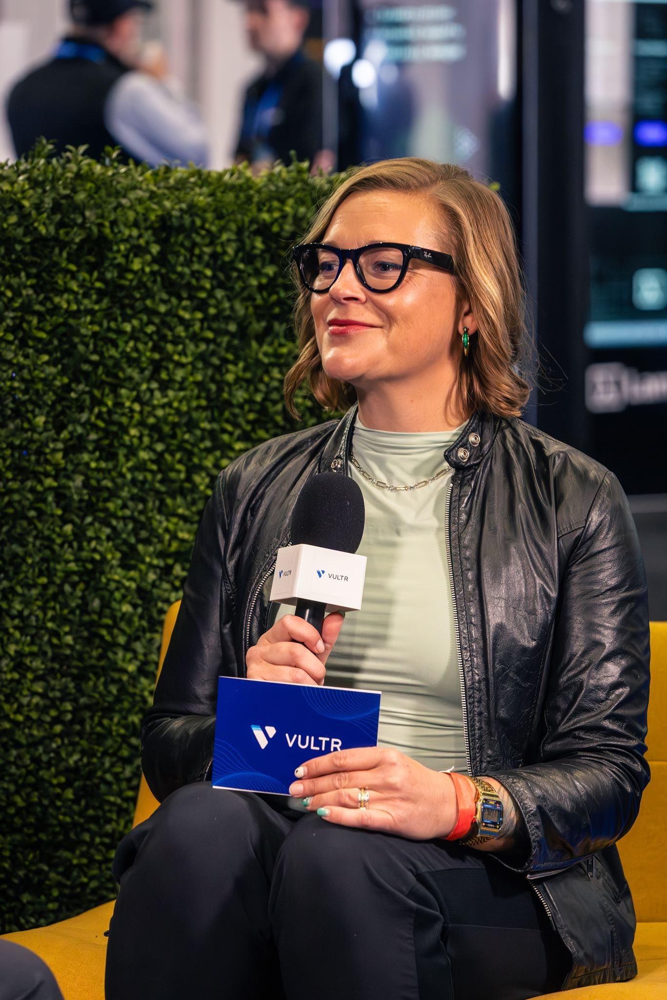 A woman with shoulder-length blonde hair, wearing glasses, a black leather jacket, and a green top, sitting and holding a VULTR brochure and a microphone in front of a green hedge with blurred people and electronic screens in the background.