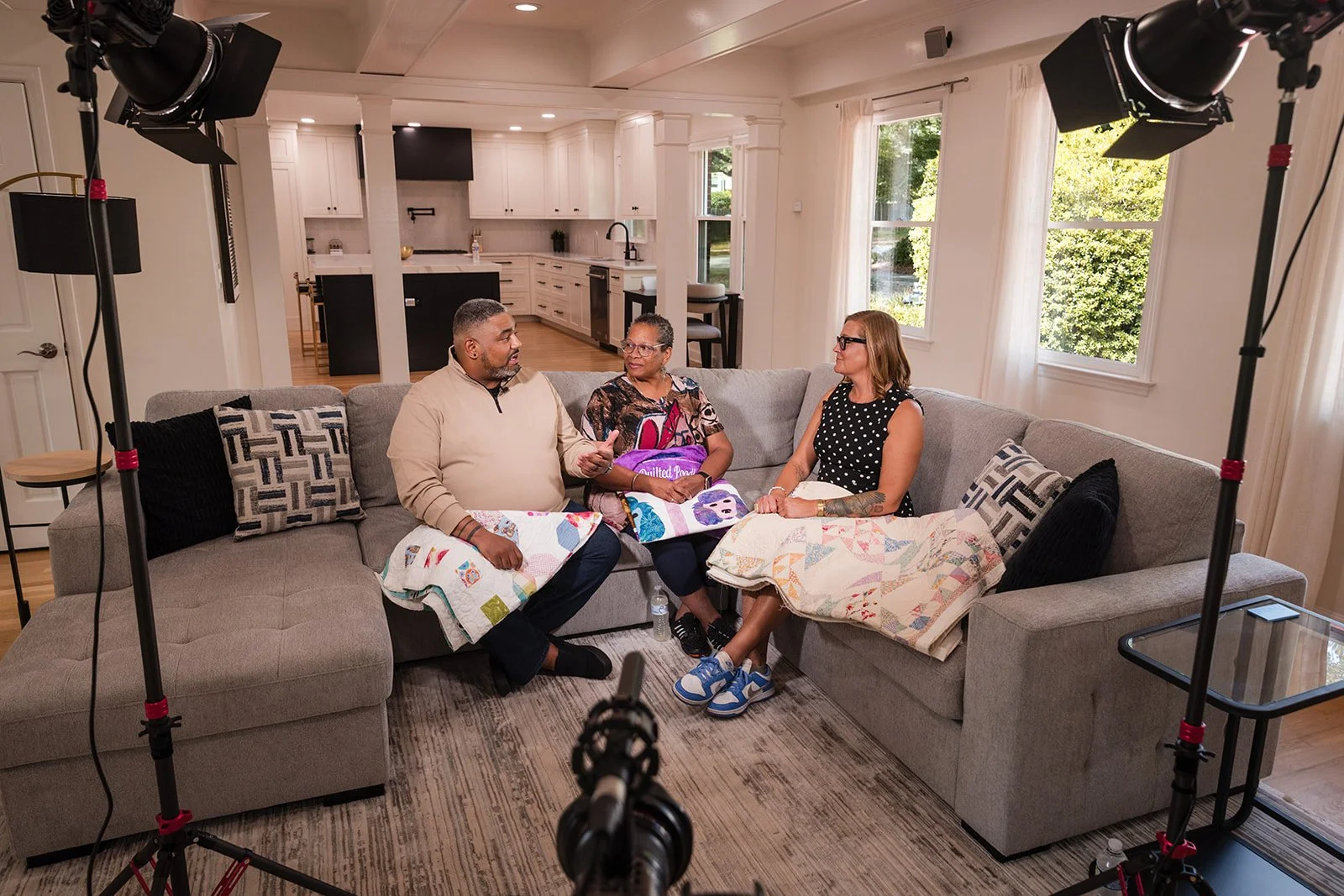 Three people sitting on a grey sectional sofa in a well-lit living room having a conversation during a recorded interview or discussion. They are using quilted blankets on their laps, and there is professional lighting equipment set up around them.