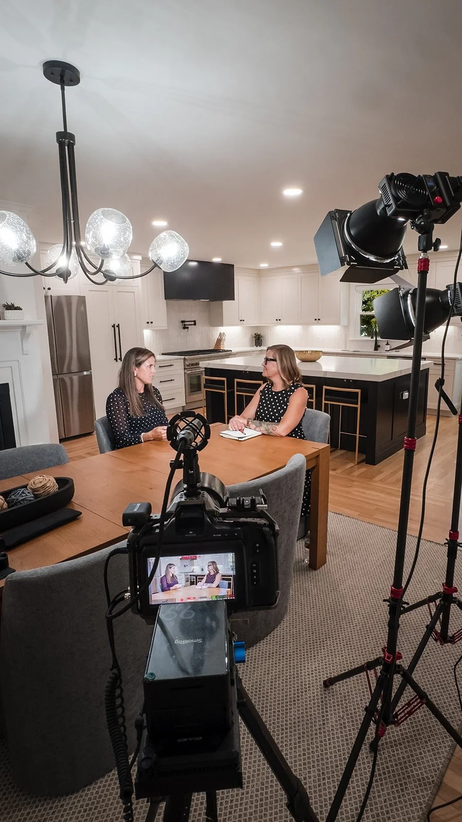Two women having a conversation during an interview in a modern kitchen, with professional camera equipment capturing the scene.