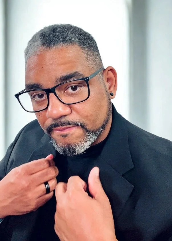 A man with short gray hair, glasses, and a beard in a black suit posing indoors.