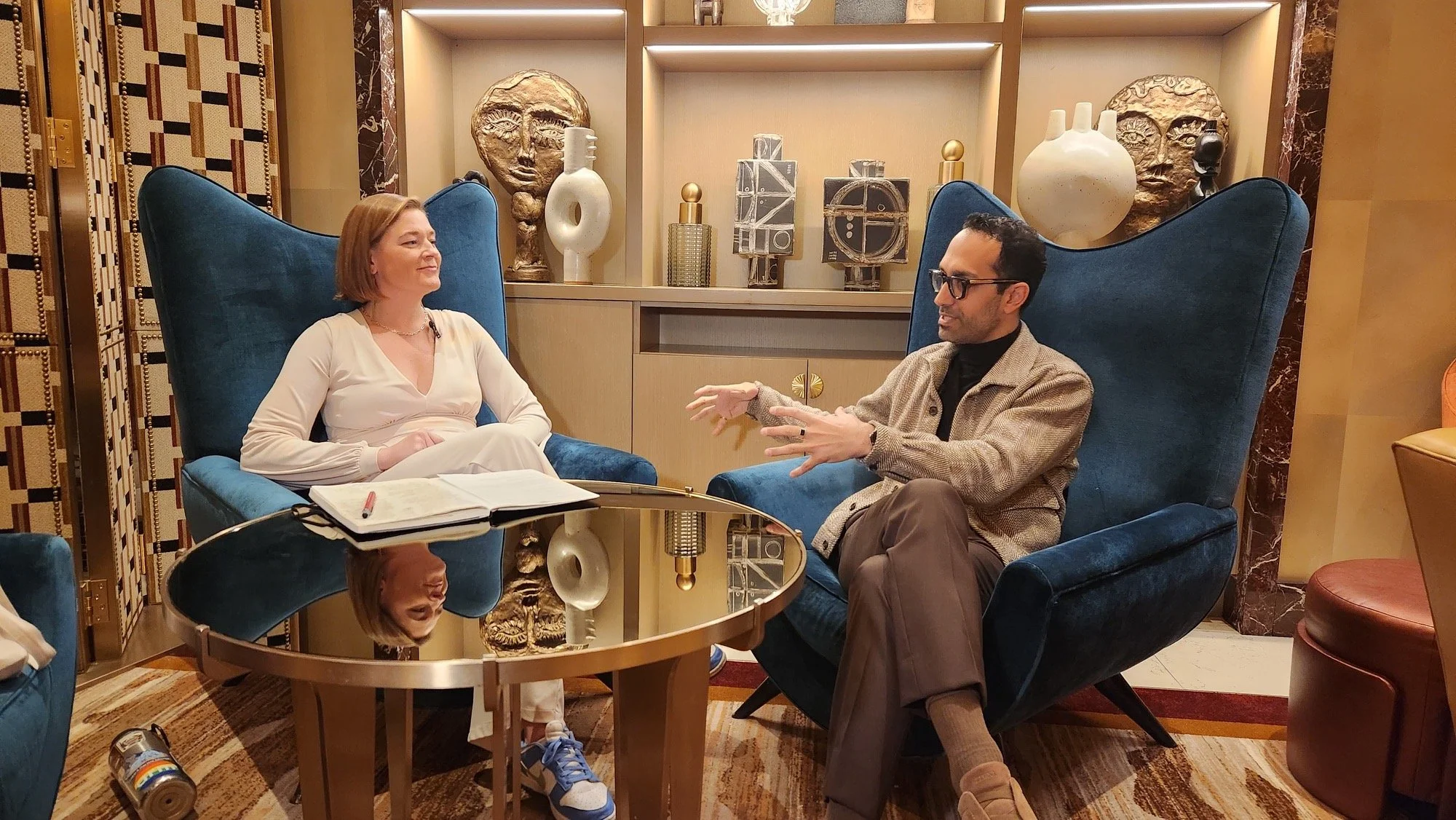 A man and a woman sit facing each other in a luxurious lounge area, engaged in conversation. The woman has shoulder-length reddish hair and is dressed in a white outfit, while the man has dark hair, glasses, and is wearing a patterned blazer. There is a round glass-top table with a notebook and a pen in front of them, and decorative art pieces, including masks and sculptures, are on the shelves behind them.
