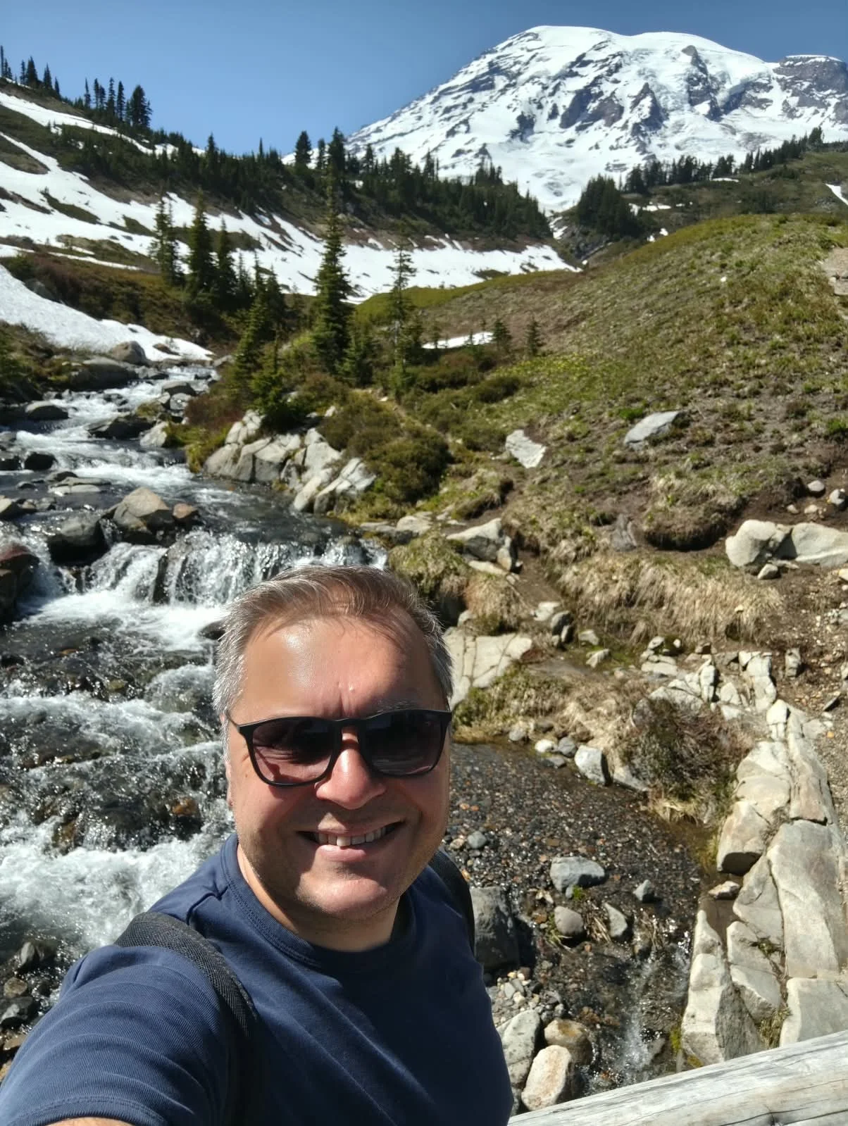 A man with sunglasses smiling in front of a mountain landscape with snow, trees, a stream, and a snow-capped peak in the background.