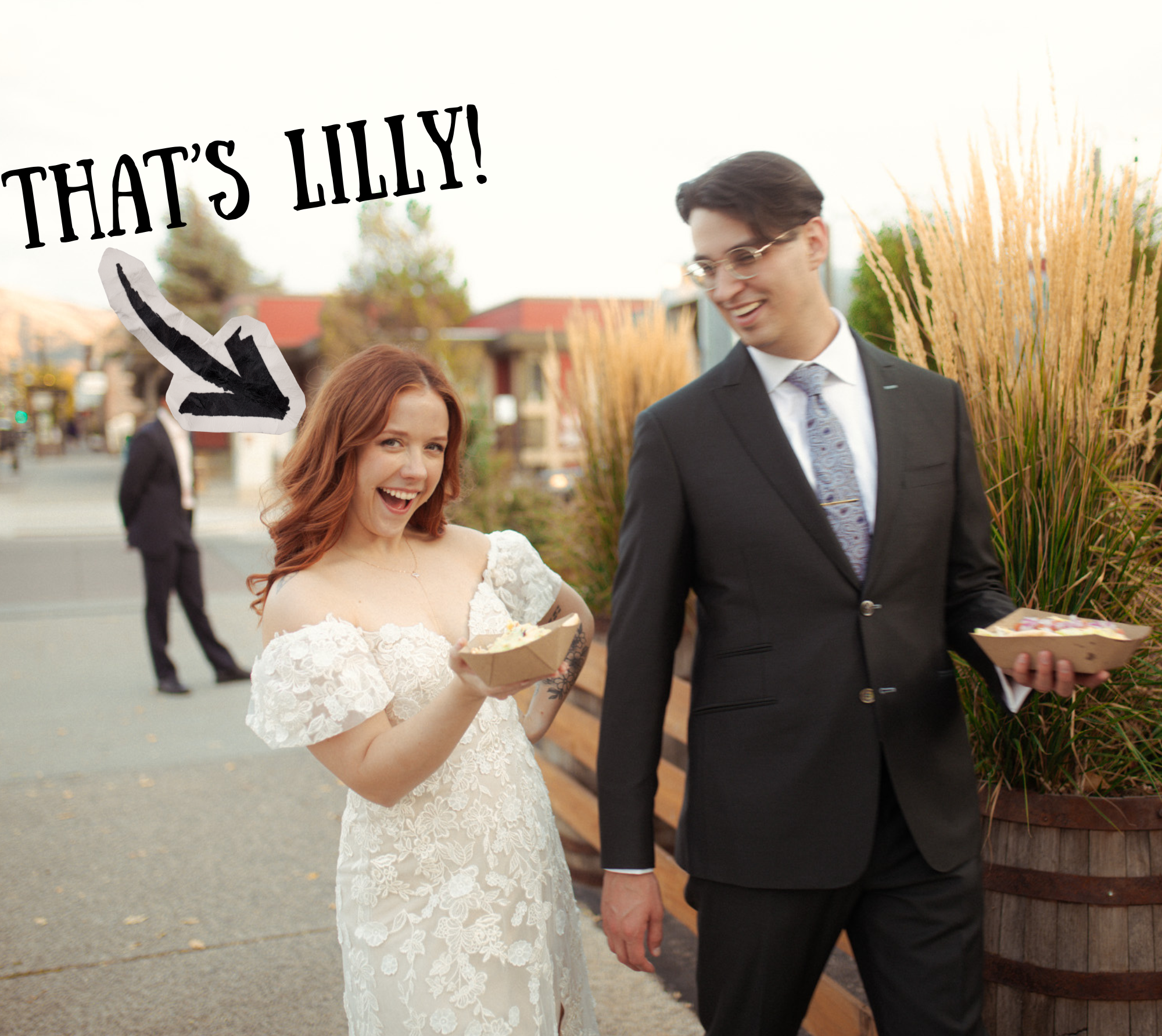 A bride in a white lace wedding dress smiling and holding a cake, walking with a groom in a dark suit and tie, holding hands, during a wedding celebration outdoors with tall ornamental grasses and a wooden fence in the background.