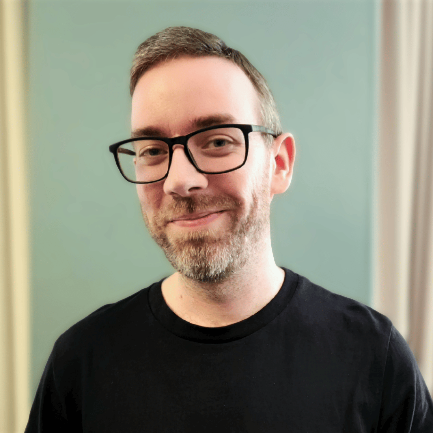 Portrait photo of author Will Irvine wearing a black t-shirt and smiling at an angle to the camera as he stands in front of a warm green coloured wall.