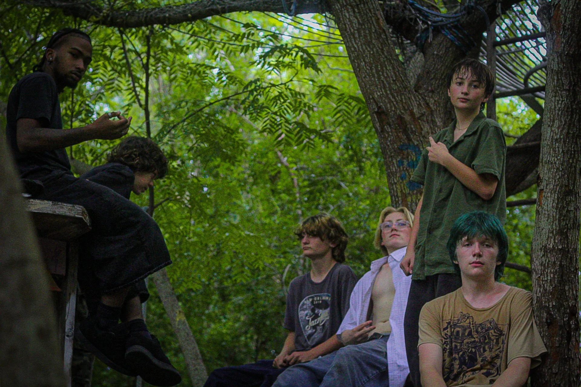Group of people in an outdoor wooded area, some sitting on a wooden platform and others standing, with trees and green leaves in the background.