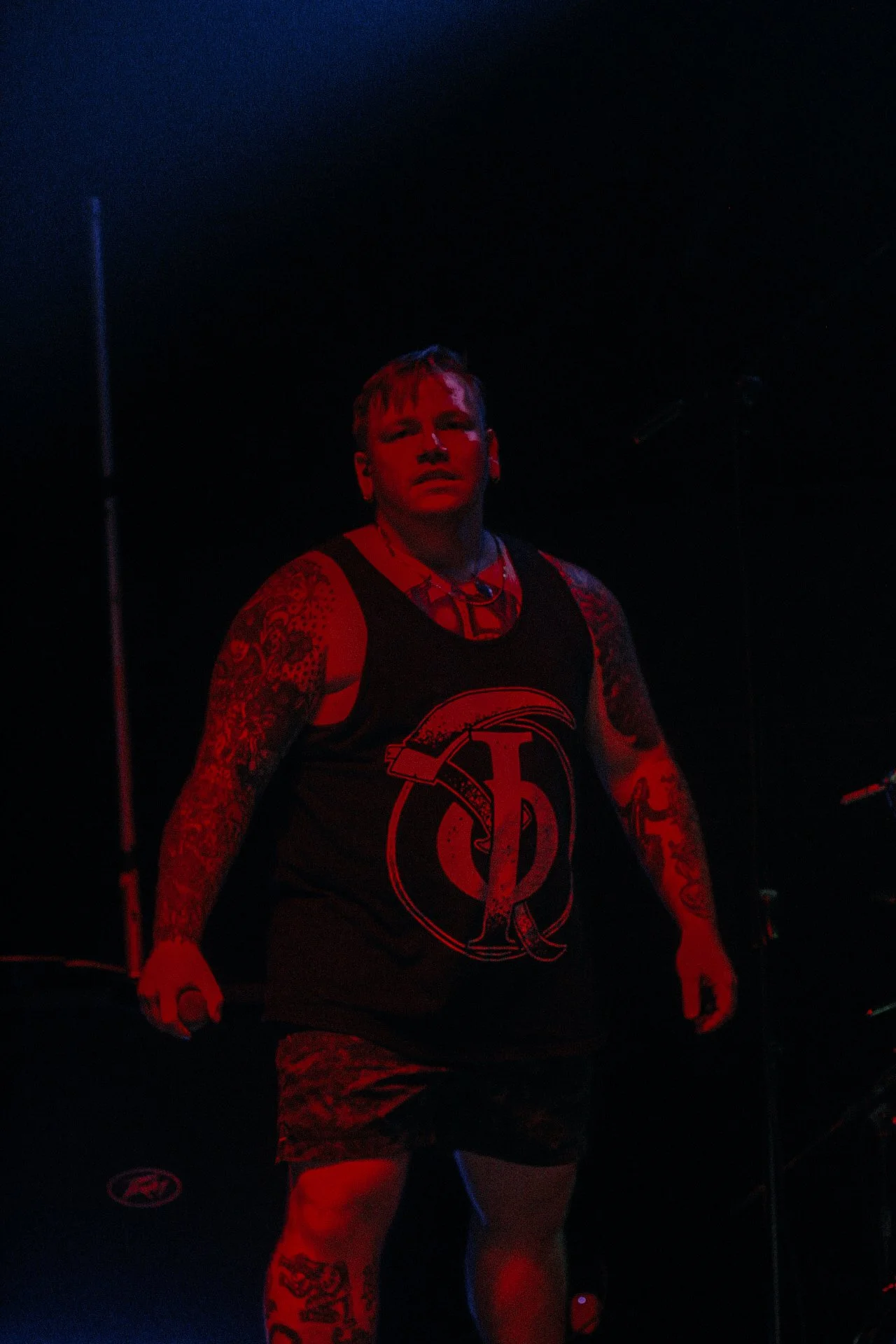A muscular man with tattoos stands in a dark environment illuminated by red lighting. He is wearing a sleeveless black shirt with a large logo on it and patterned shorts.