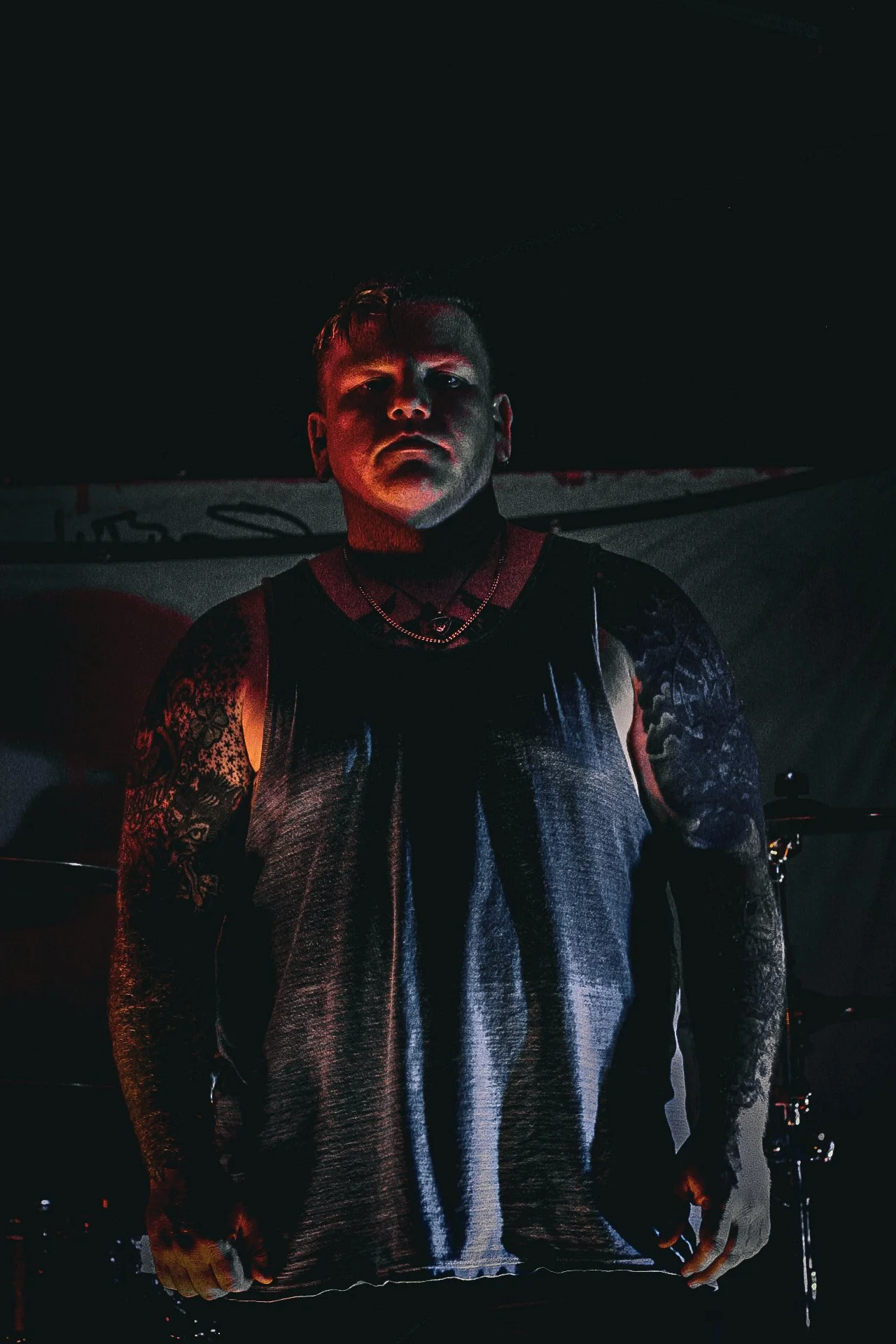 A young man with tattoos on his arms, wearing a sleeveless top and necklace, standing in a dark room with dim lighting and a drum set in the background.