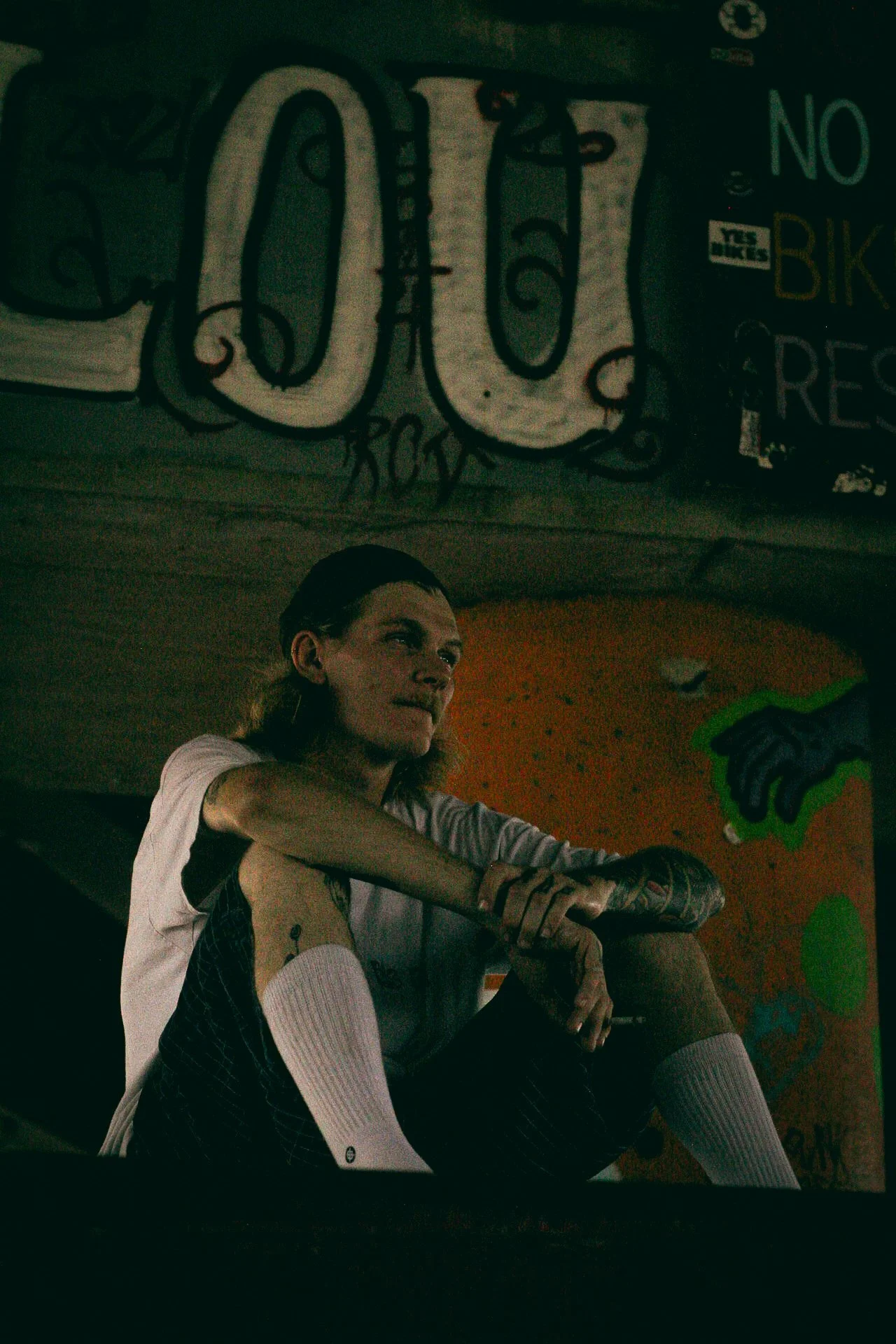 A person with long hair seated with legs crossed, wearing a white and black shirt and pink knee-high socks, in a dimly lit room with colorful graffiti and street art on the walls.