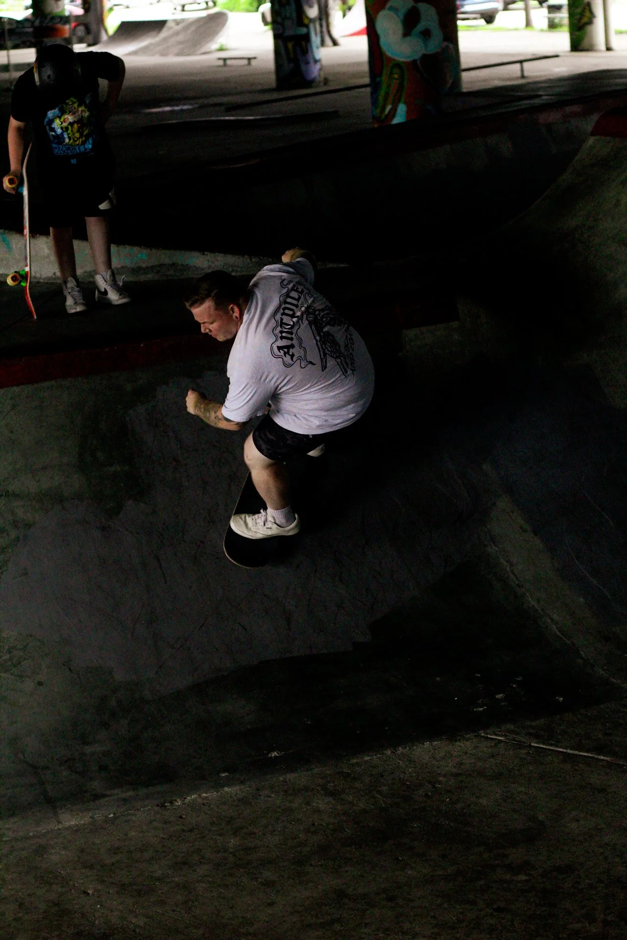 A person doing a skateboarding trick in a skatepark, with another person standing nearby holding a skateboard.