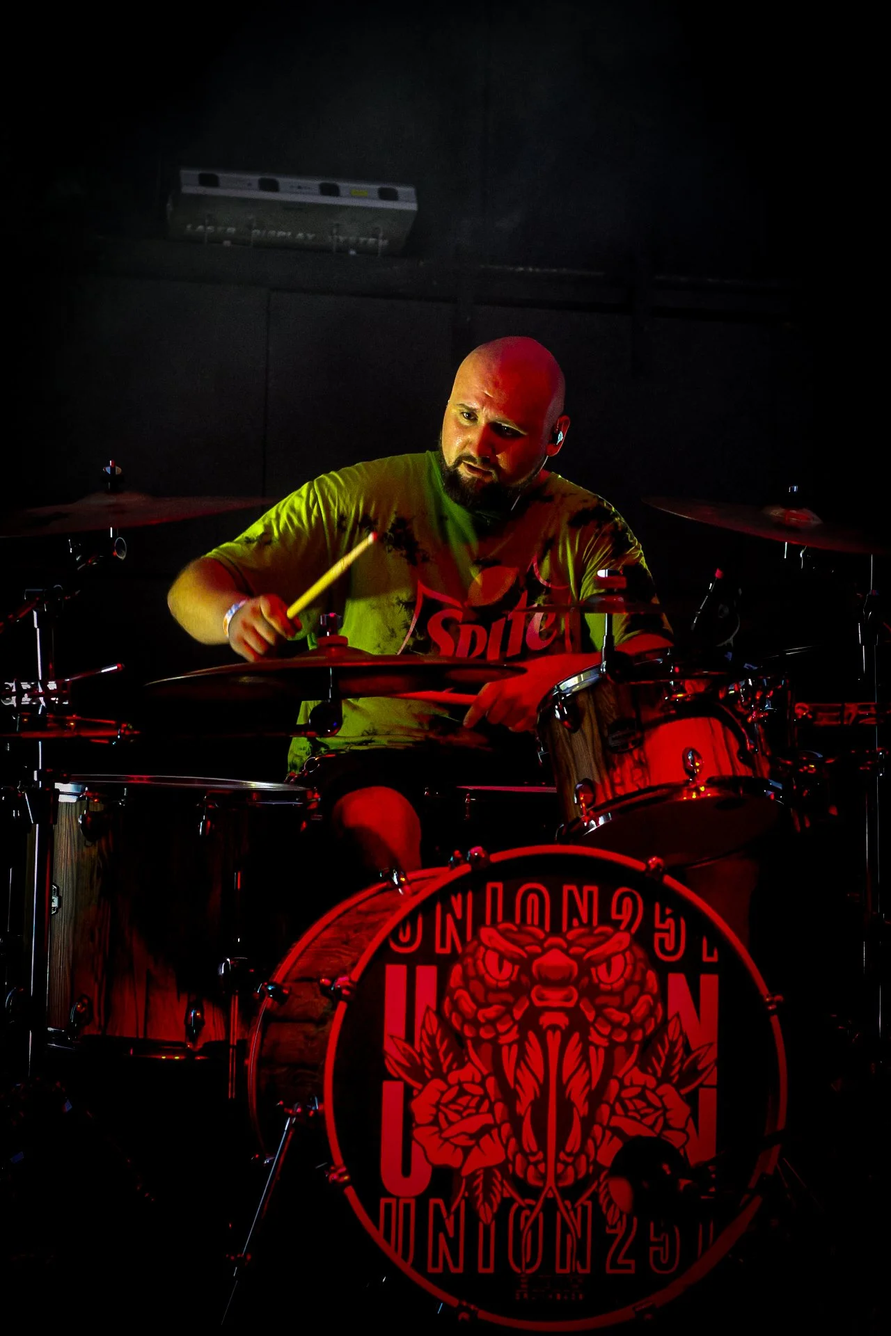 A man playing drums on stage, illuminated by colored lights, with a drum kit featuring a logo of a tiger and text that reads 'LONDON 25'.