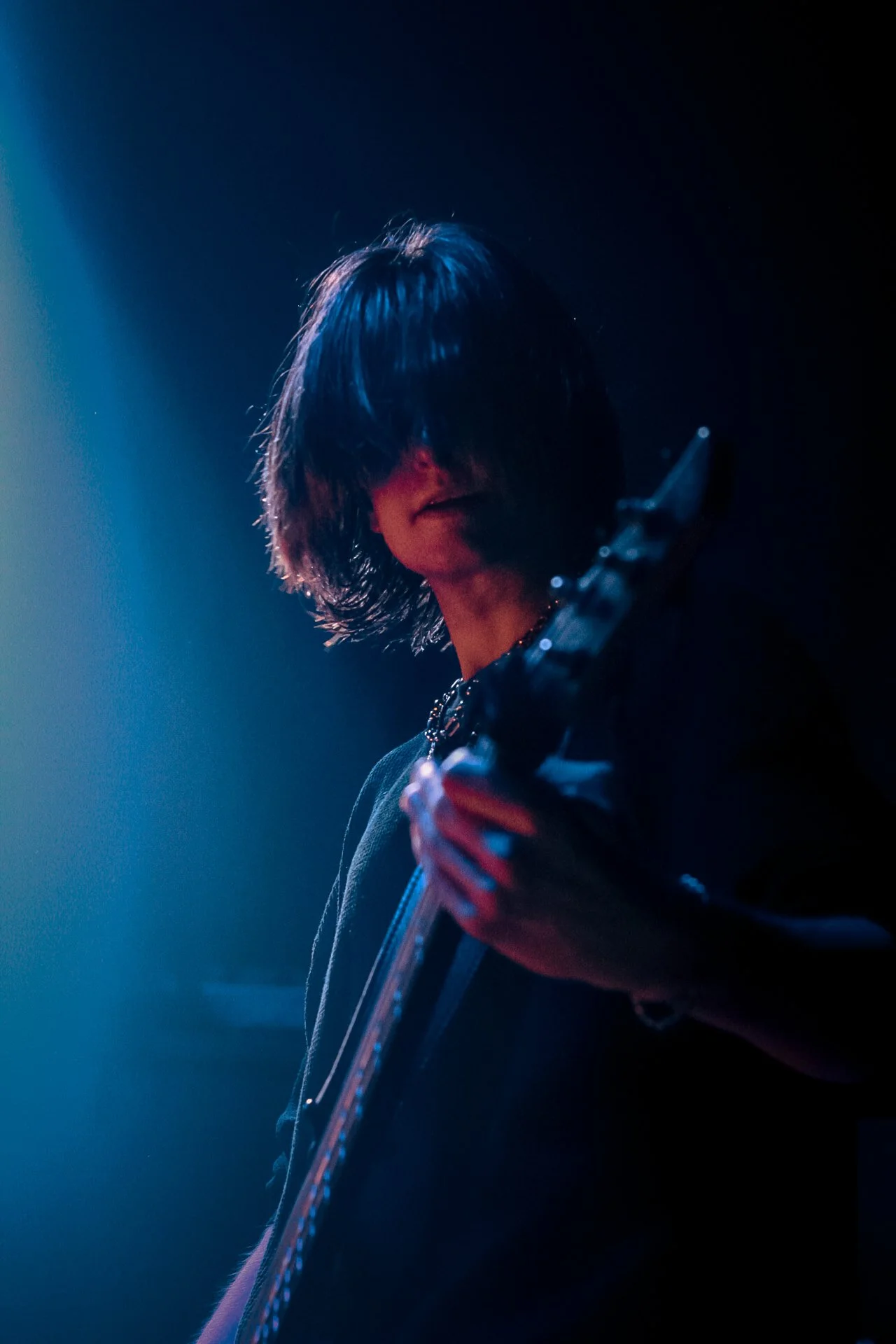 A person with long dark hair playing an electric guitar on stage, illuminated by blue and purple lighting.