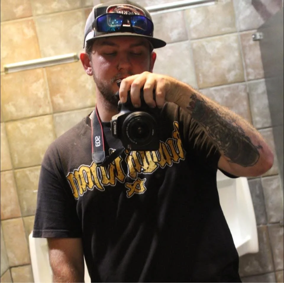 A man taking a selfie in a bathroom mirror with a camera. He is wearing a black T-shirt with gold lettering, a baseball cap, and sunglasses on the cap's brim. He has tattoos on his right arm and is standing in front of beige and gray tiled walls.
