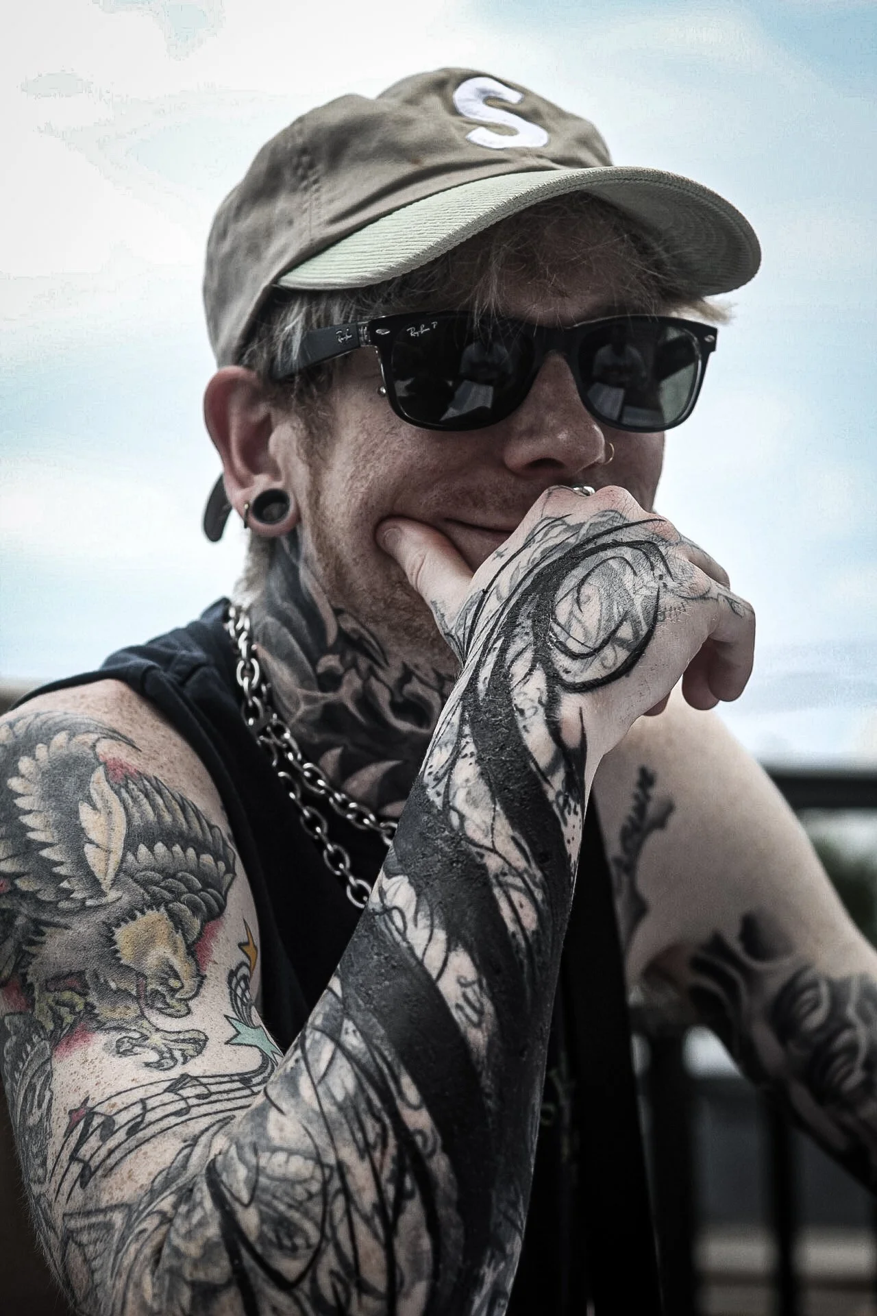 A person with tattoos on their arms and neck, wearing sunglasses, a beige cap, and a black tank top, sitting outdoors.