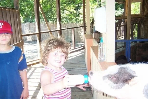 A young girl with curly hair, wearing a striped pink and white tank top, feeding a pig with a bottle on a farm or petting zoo deck. A boy in a blue shirt and red cap stands nearby. The setting is outdoors with wooden fencing and trees in the background.