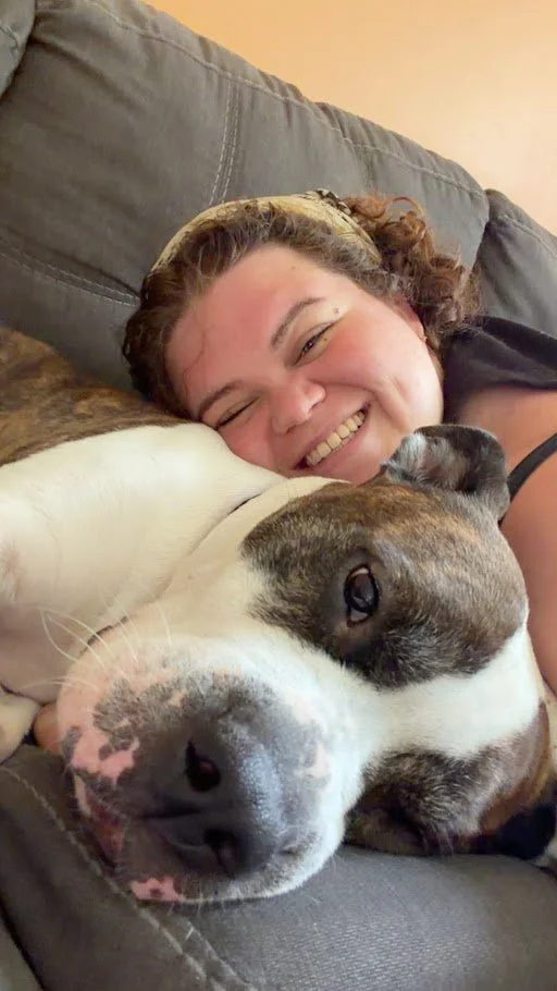 A woman lying on a couch with a big, relaxed dog and a small puppy, all smiling and close together.