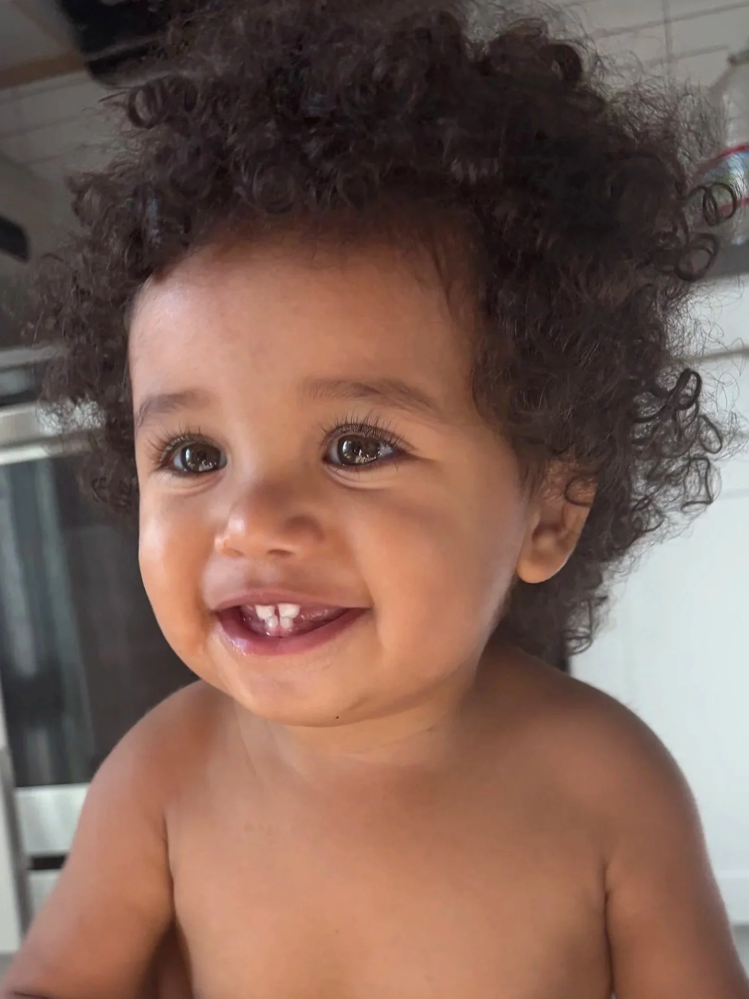 Close-up of a young child with curly dark hair, smiling, showing some missing front teeth, and with expressive brown eyes.