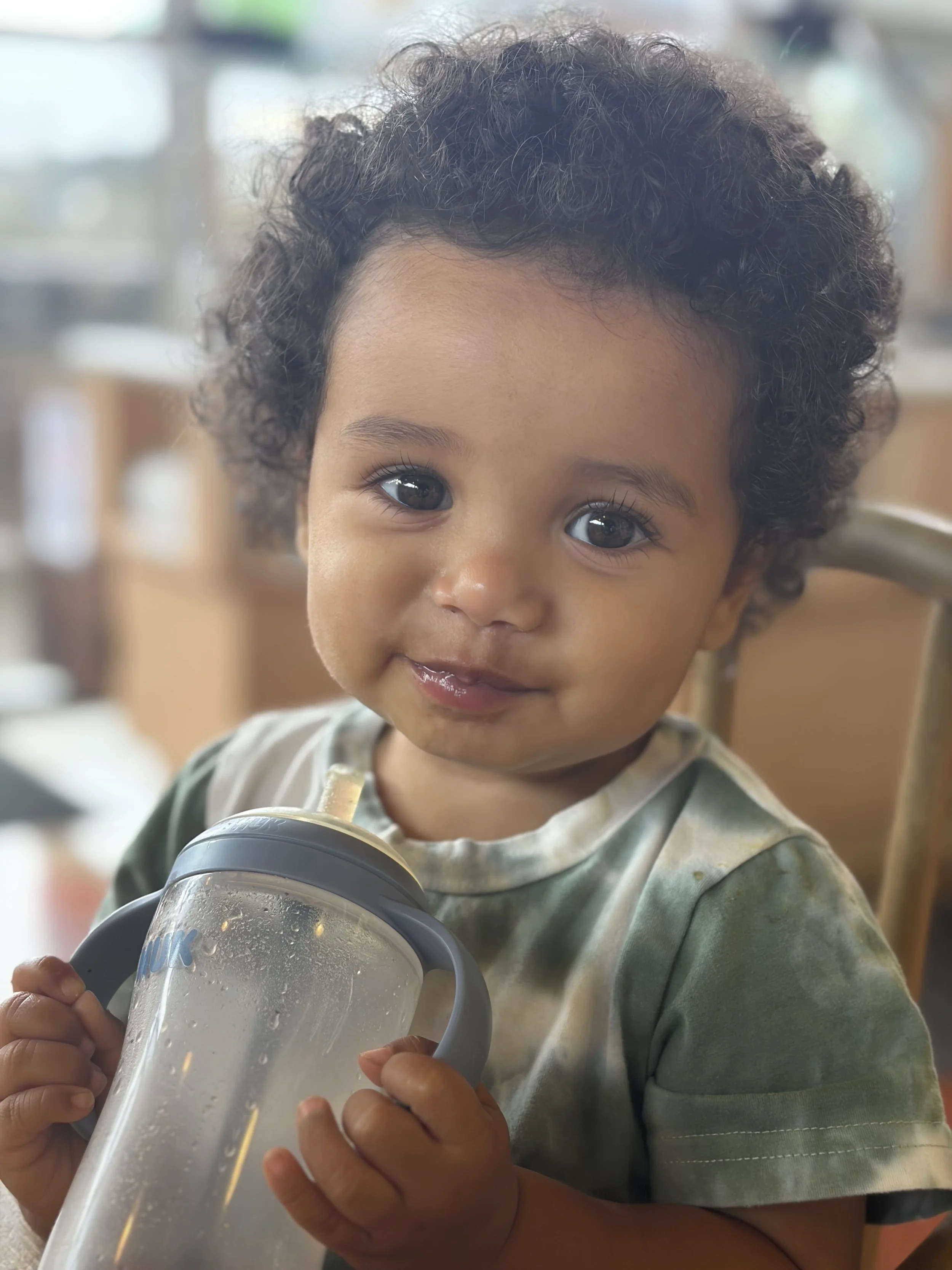 A young child with curly hair smiling while holding a large clear cup with a straw, filled with a cold beverage, in a cozy indoor setting.