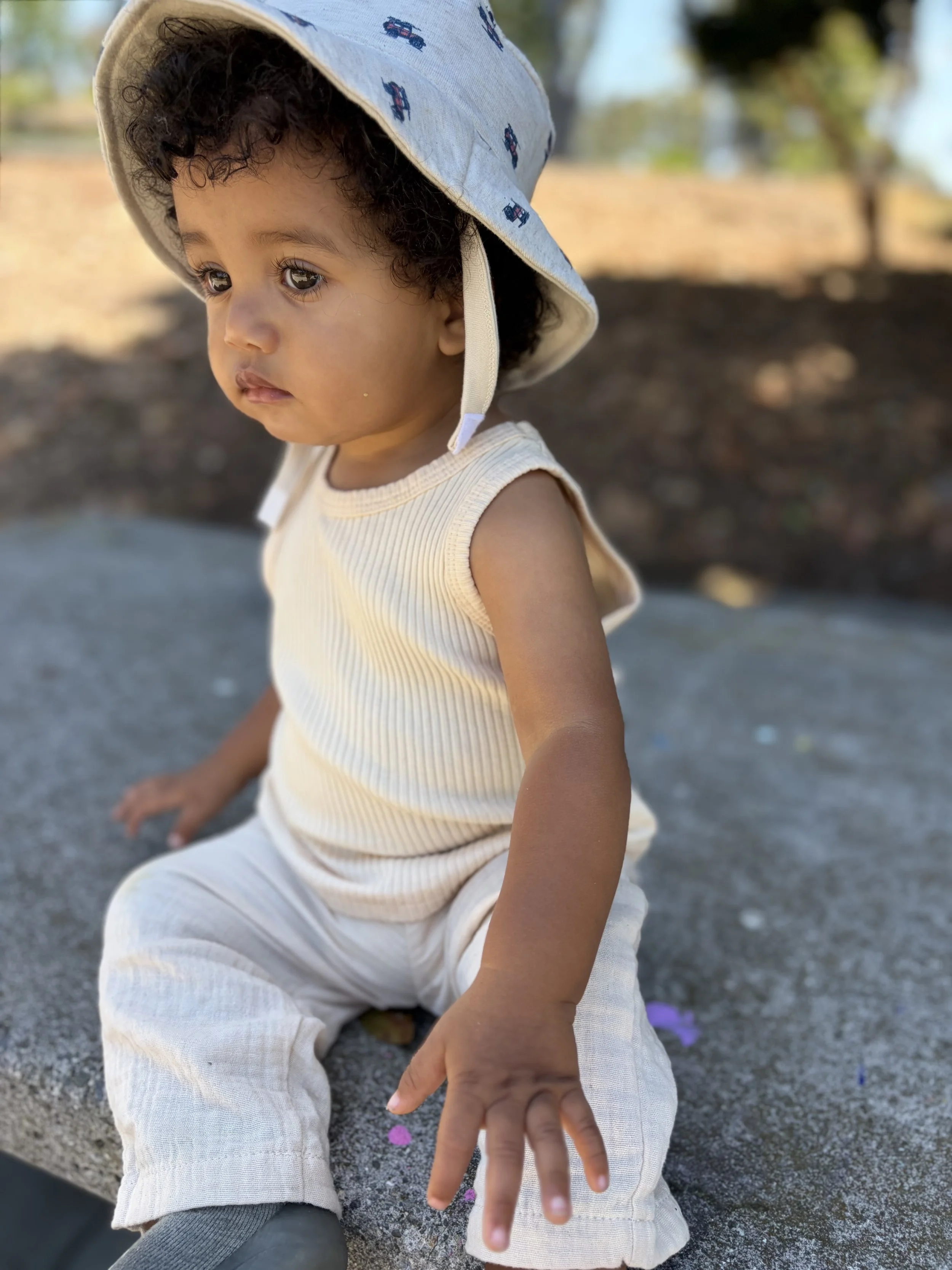 A young child with curly hair wearing a sun hat and light-colored clothing, sitting on a concrete surface outdoors, reaching out with their hand and playing with pink and purple flower petals scattered on the ground.