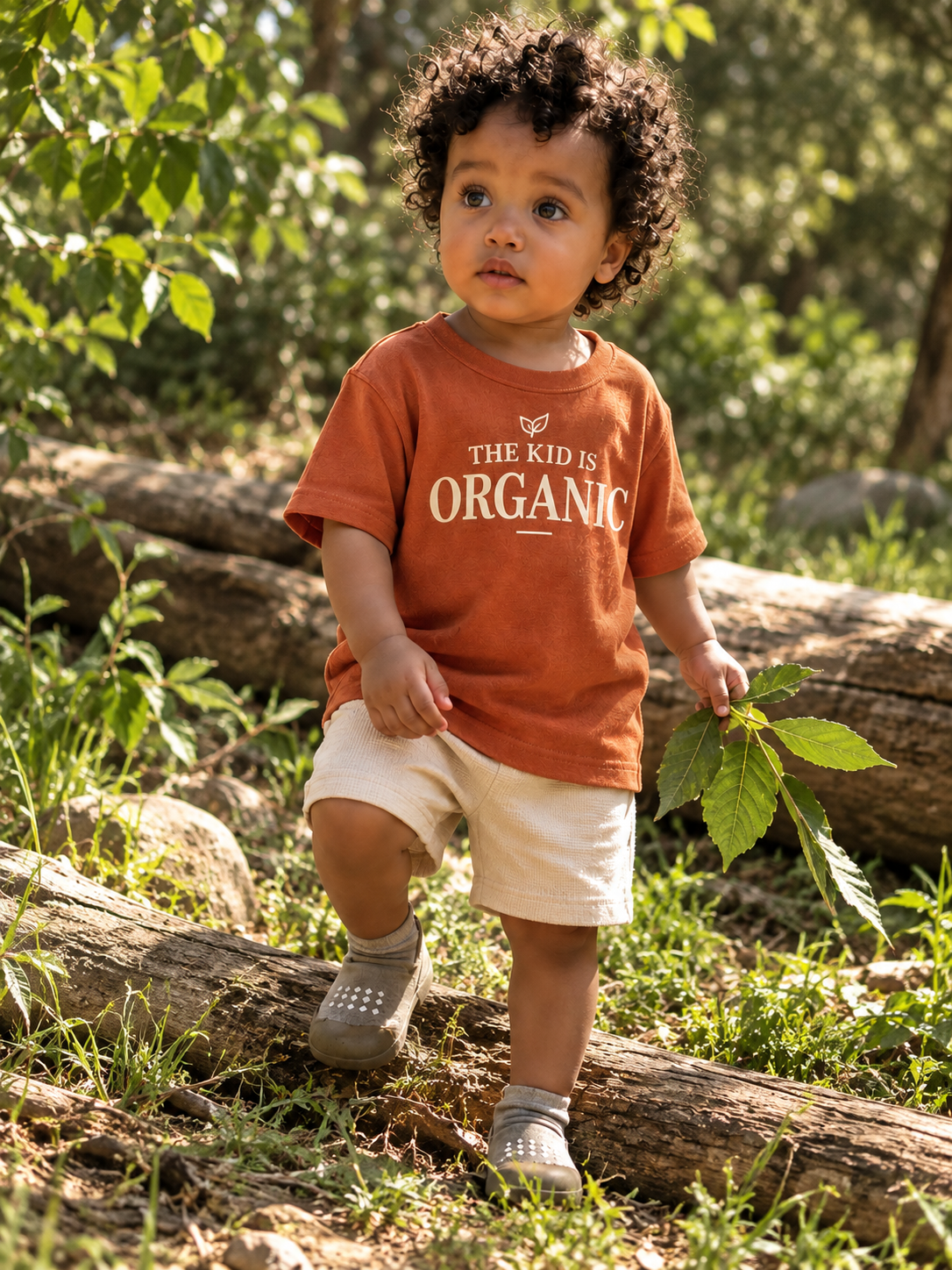 Young child walking outdoors on a fallen log, holding a green leaf, wearing a burnt orange T-shirt with the words 'The Kid is Organic', beige shorts, and gray shoes, surrounded by greenery and dappled sunlight.